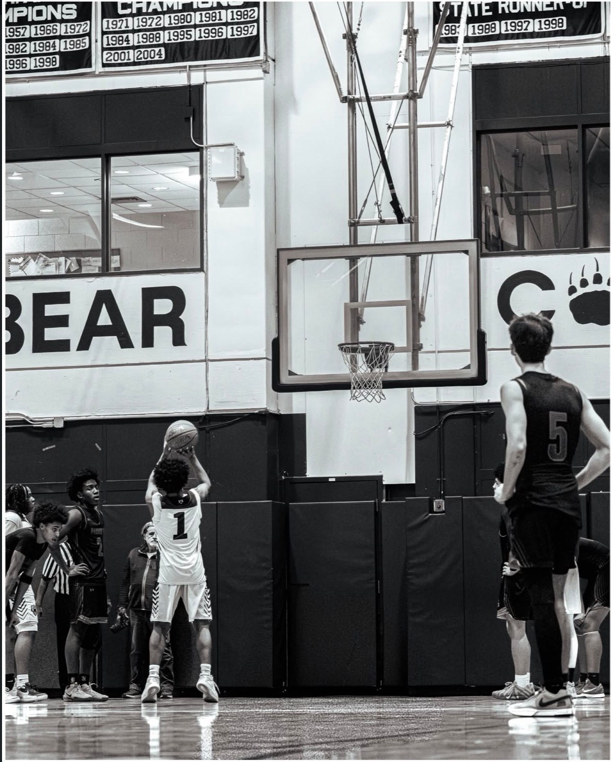 Black and white basketball photo showing players side by side, the hoop during a free throw indoors. 