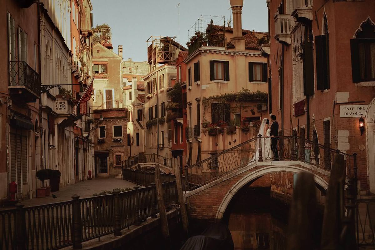 Daisy and Luis pause to watch the sun flare above Venice from their vantage point above a canal.