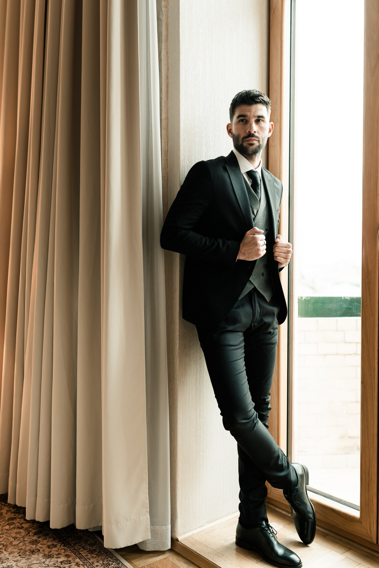 Mladoženja u crnom odelu naslonjen stoji pored prozora, gledajući u stranu u elegantnom i mirnom portretu.
