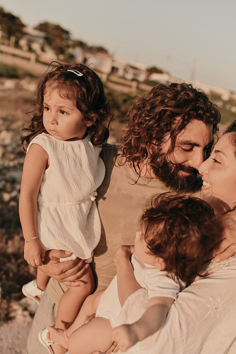 A family with two children shares a tender moment. Dad holds the older daughter, mom holds the baby. They smile in the warm sunset light.
