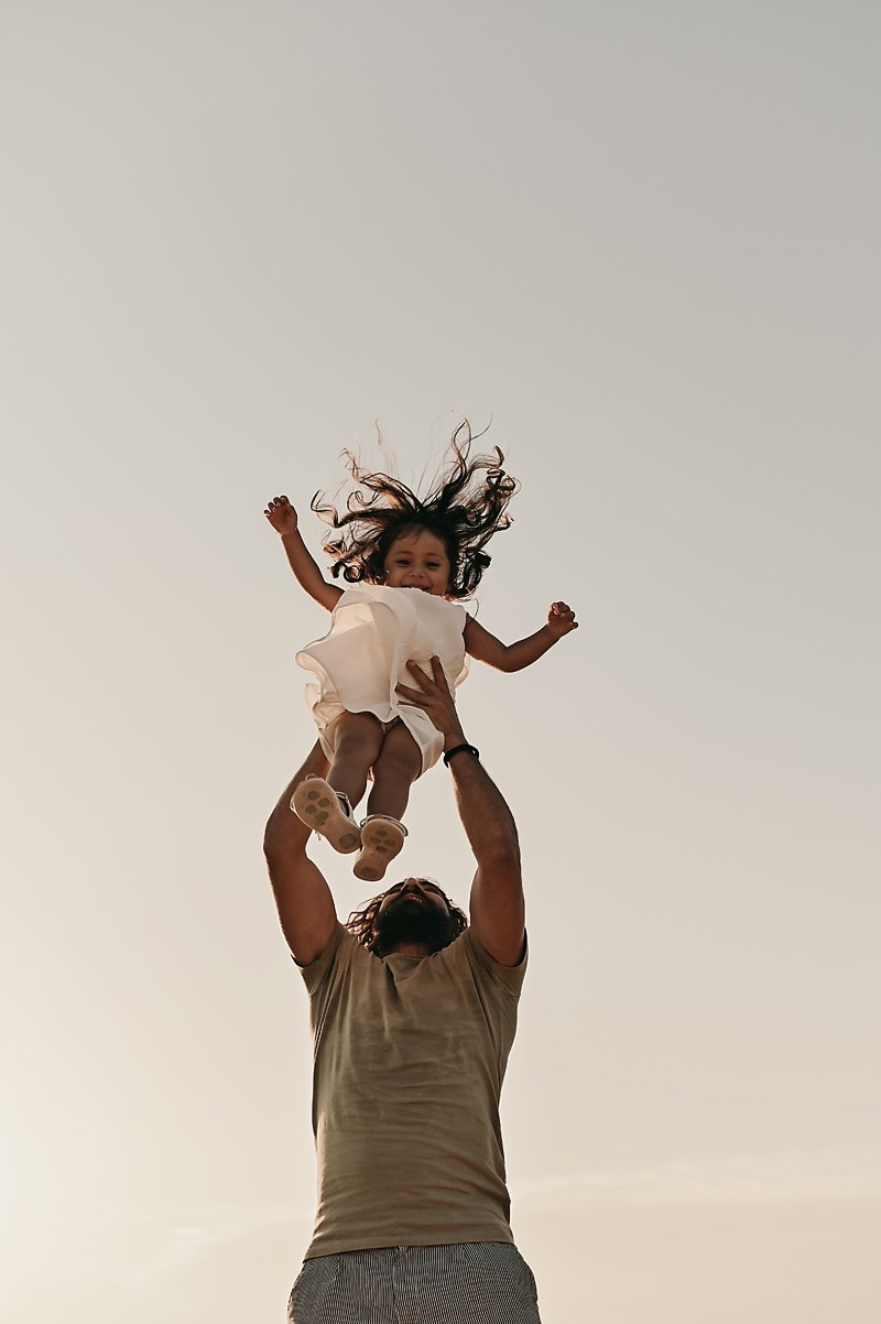 A father tosses his daughter in the air at sunset. Her hair flows freely as she smiles against the sky.