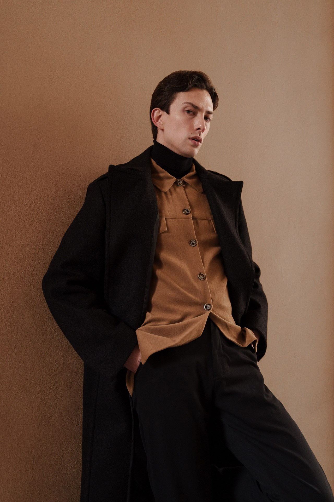 Male model leaning against textured wall in black coat and camel shirt