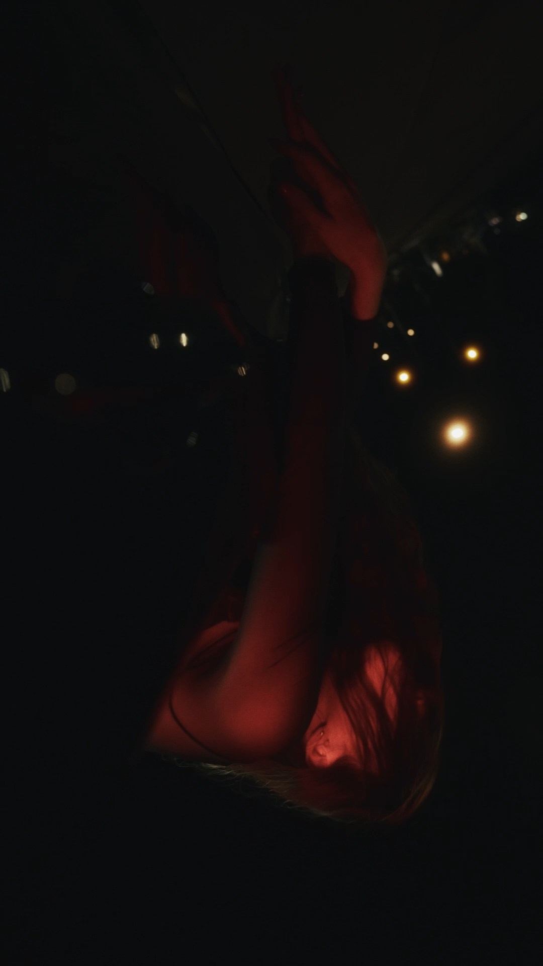 A close-up of a person reclining on a surface, illuminated by a mix of red and ambient city lighting, with their face turned upwards in a dramatic and reflective pose.