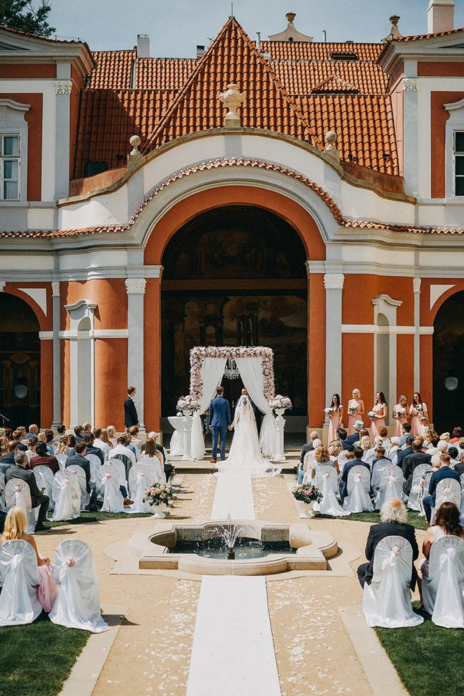 A bride and groom stand under a floral and chandelier altar piece especially created for their Ledebour Garden outdoor wedding in Prague.
