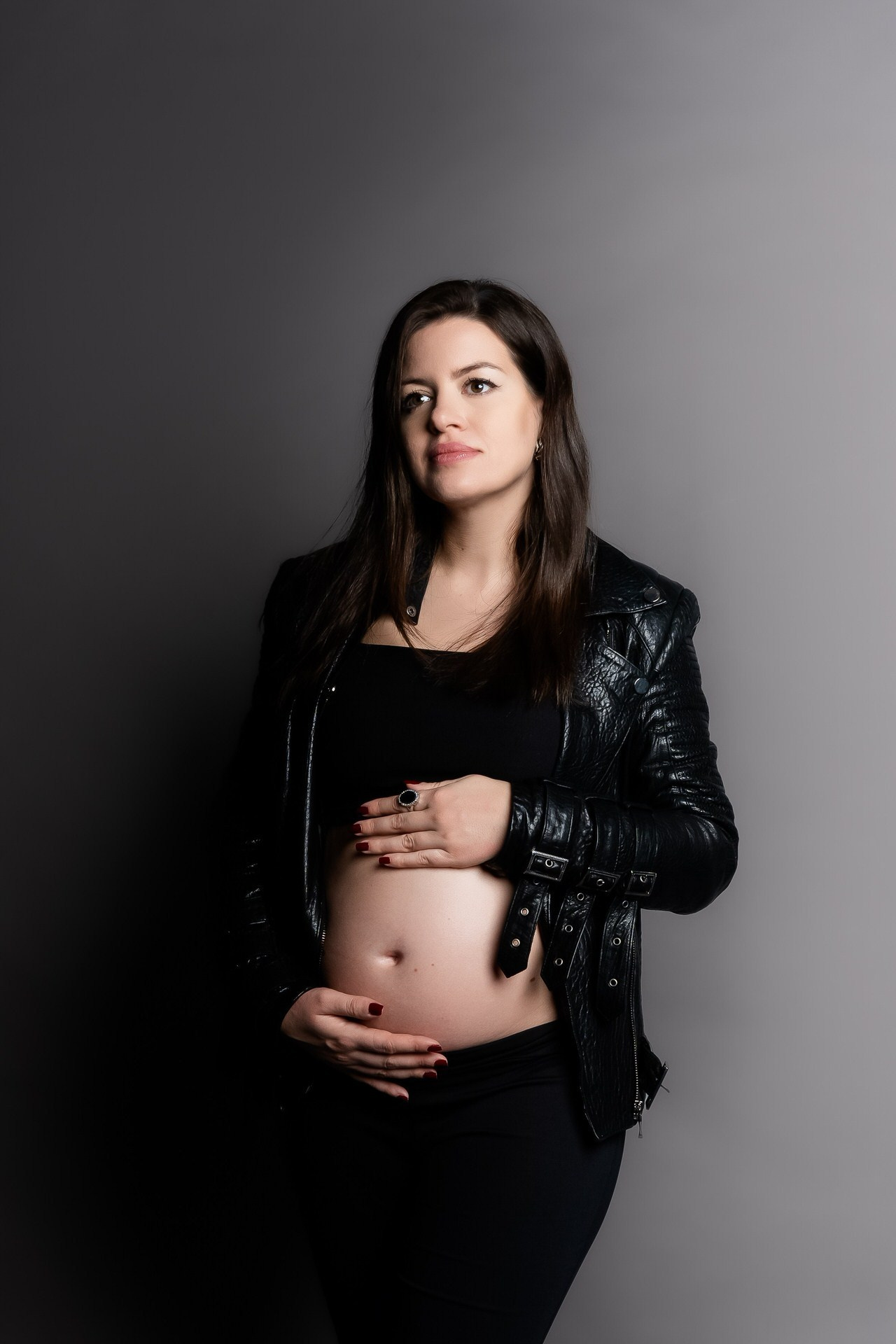 Portret trudnice u crnoj odeći i kožnoj jakni, koja nežno drži stomak pred tamnosivom studijskom pozadinom.