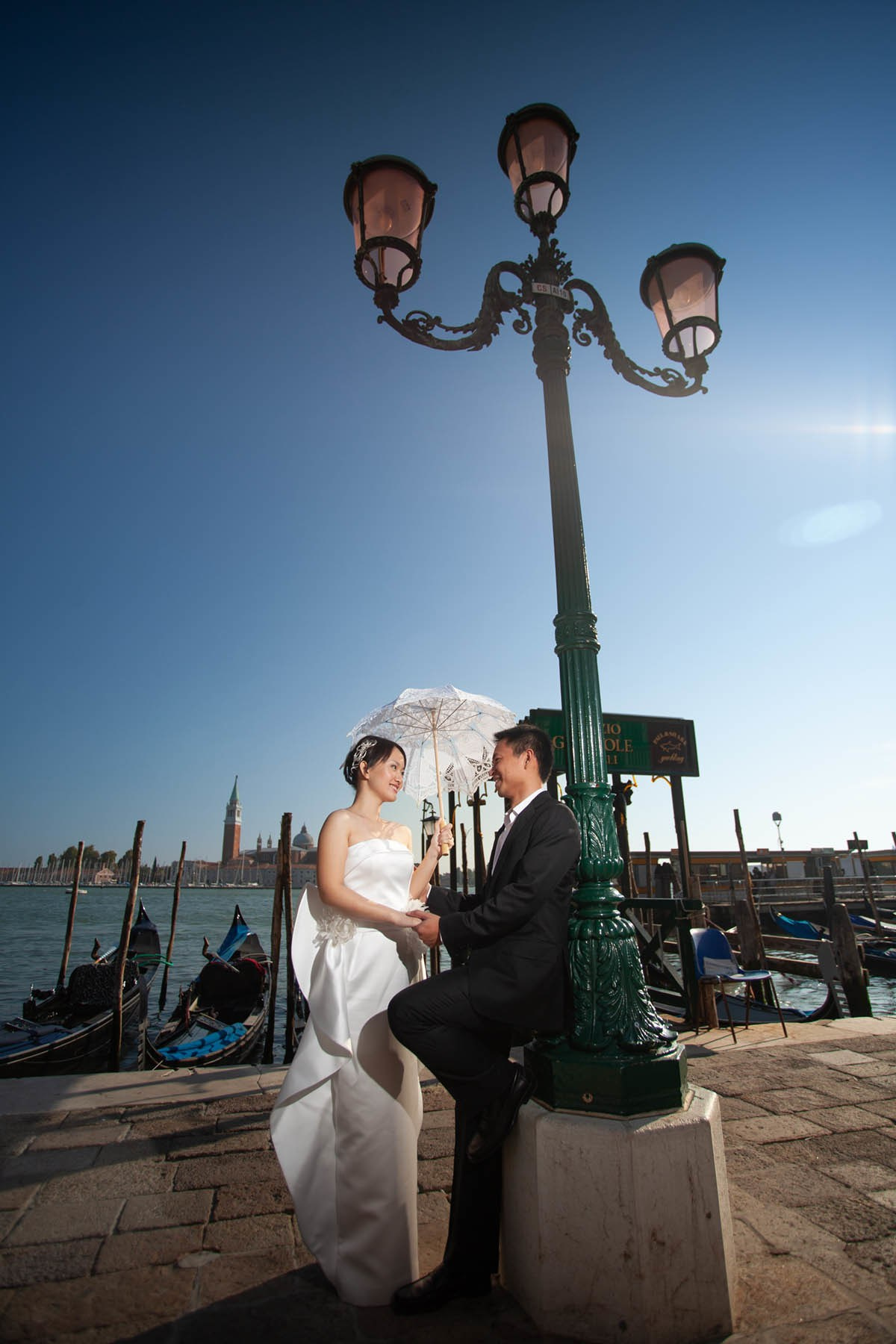 Beaming Thai bride with parasol chatting with partner near gondola on Venice canal.