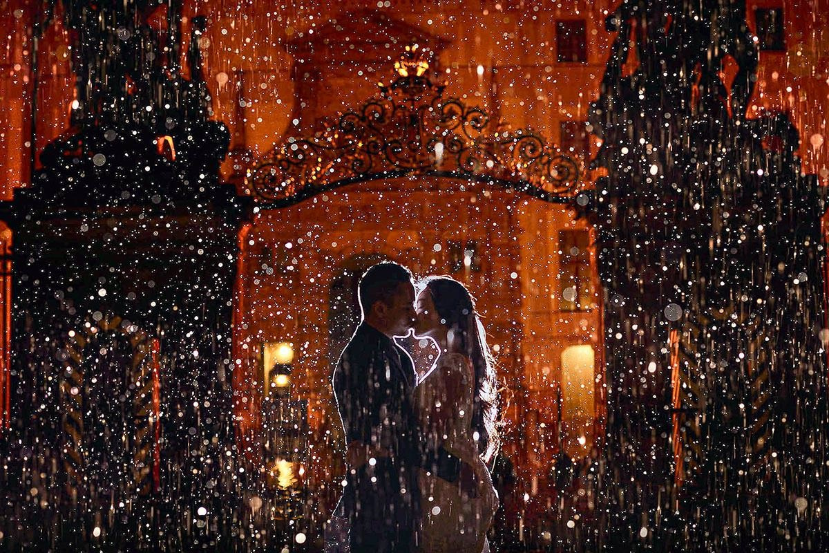 Raindrops are frozen in backlight as a young couple kisses in front of Prague Castle guardshacks at night.