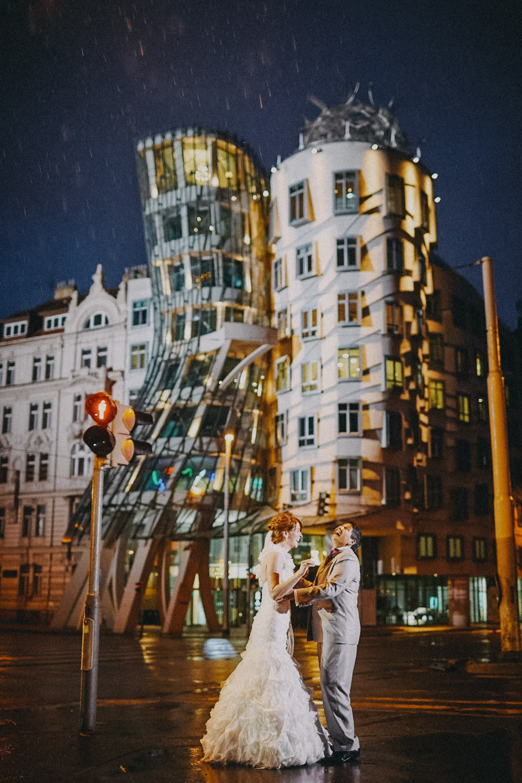 Same sex couple bride laughing as they embrace in the rain in front of the iconic Dancing House, or Ginger & Fred in Prague.
