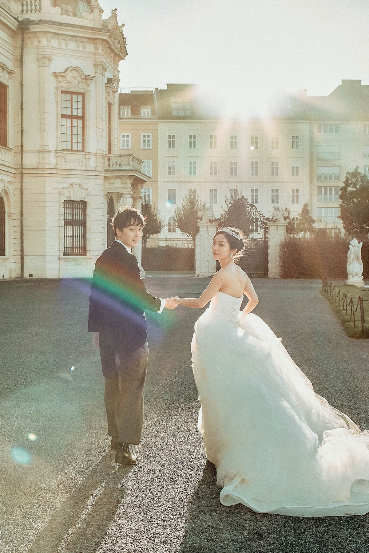 Asian newlyweds look back towards the camera as they walk in the sunlight, at the Belvedere Palace in Vienna. 