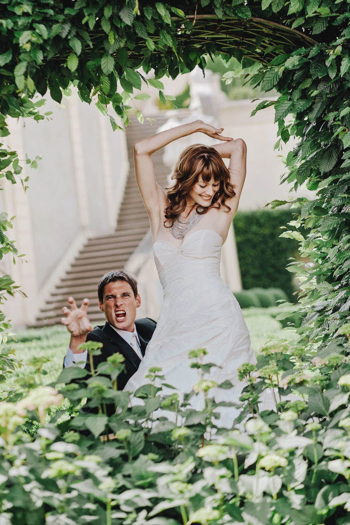 A groom makes a tiger gesture as his beautiful red-haired bride strikes a sexy pose during their Vrtba Garden summer wedding in Prague.