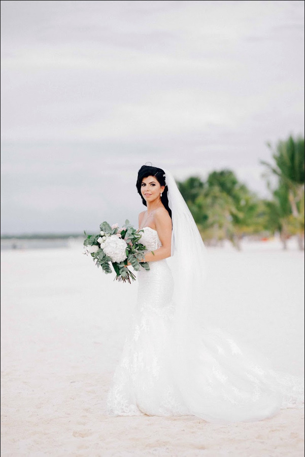 Невеста позирует фотографу на пляже возле пляжного ресторана, она одета в классическое белое платье и фату, в руках у нее красивый свадебный букет. 