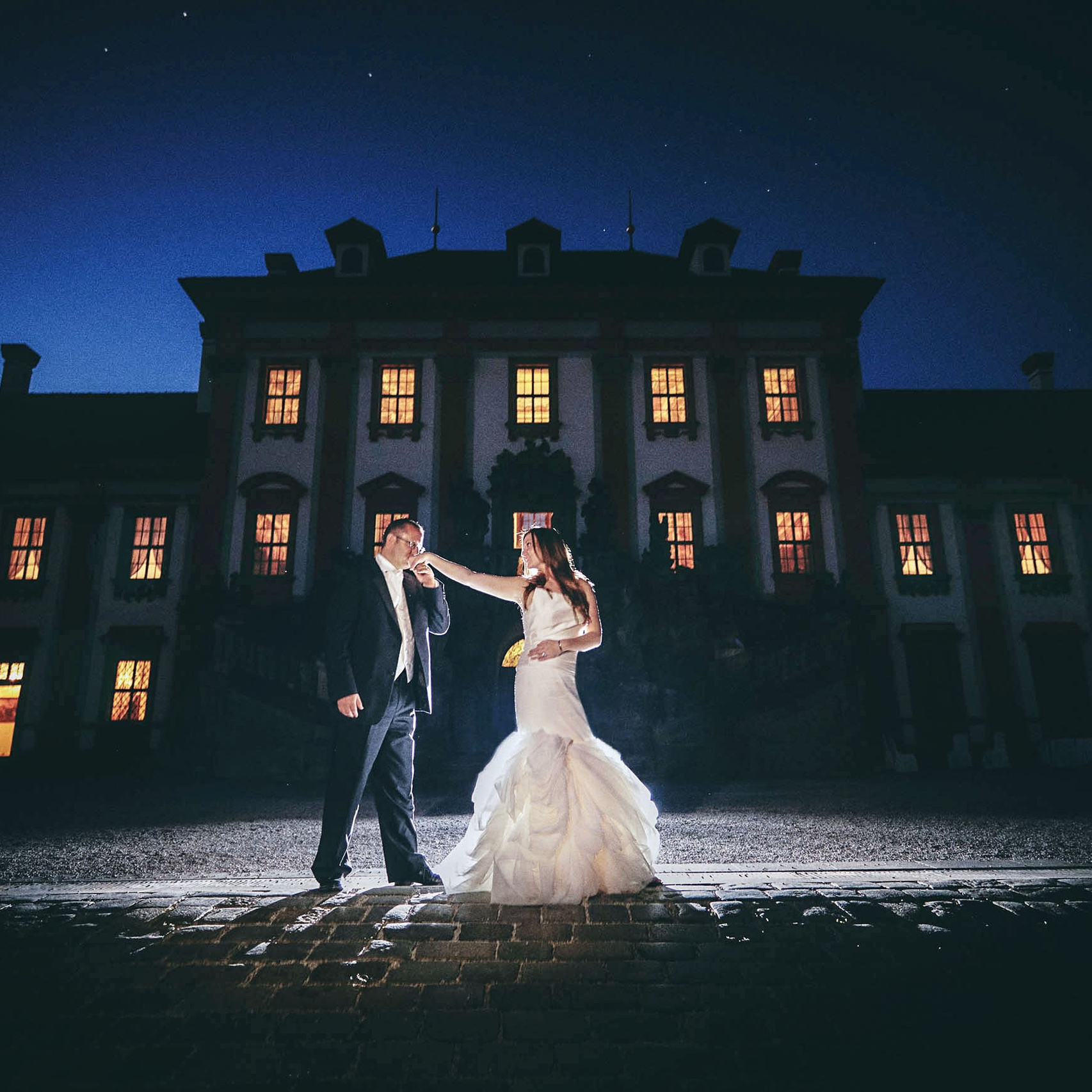 A grom kisses the hand of his stylishly dressed bride against the night time sky above the historic and opulent Troja Castle in Prague.