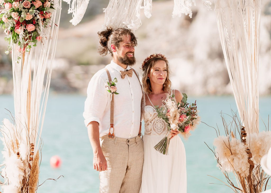 Foto einer Hochzeit in Lindos: Ein Paar steht unter einem geschmückten Makramee-Bogen. Der Bräutigam in beiger Leinenkleidung, die Braut im Spitzenkleid mit Blumenkranz und Brautstrauß. Der Bogen ist mit rosa und beigen Blumen sowie Pampasgras dekoriert. Das türkisfarbene Meer bildet den Hintergrund.