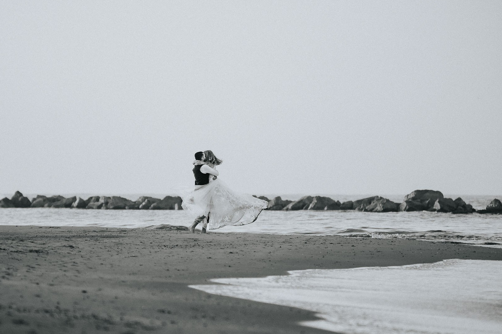 Sposi in un romantico abbraccio sulla spiaggia deserta, con la sposa sollevata in un elegante abito bianco e lo sposo in abito formale, entrambi immersi in un'atmosfera tranquilla e amorosa, catturati in una scena in bianco e nero.