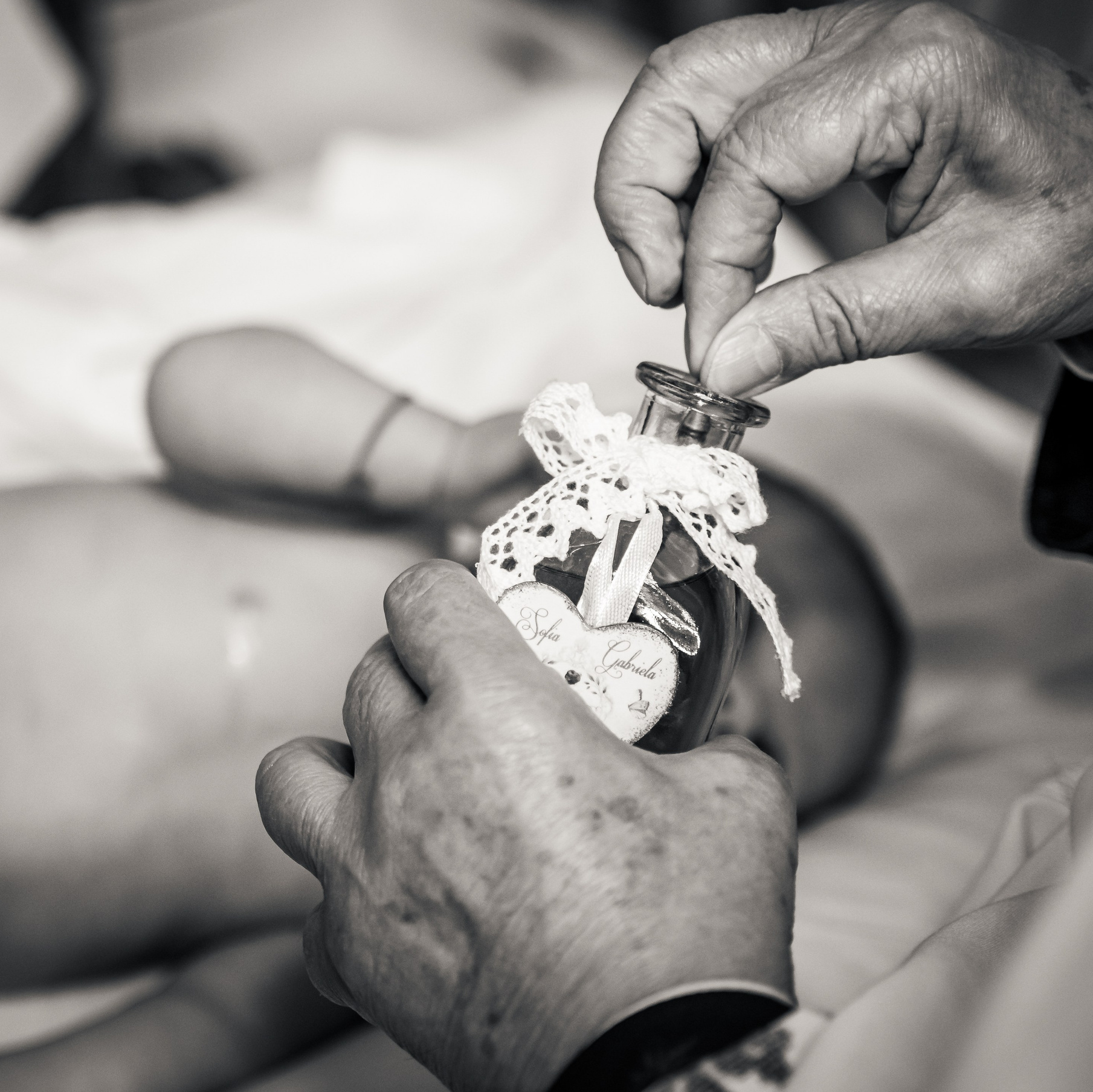 Detaliu alb-negru la un botez în București, mâinile preotului ținând sticla cu mir deasupra bebelușului, incadru.ro.