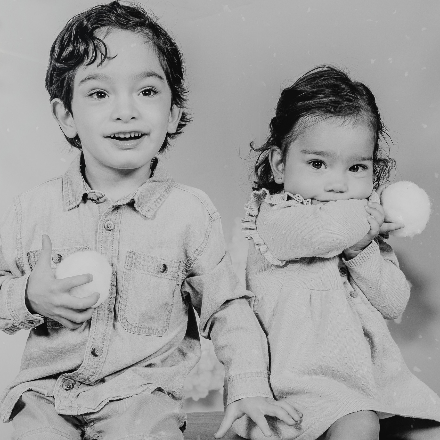 Dos niños posando en una sesión de fotos navideña, vestidos con ropa festiva y rodeados de decoraciones tradicionales como luces, árboles de Navidad y adornos