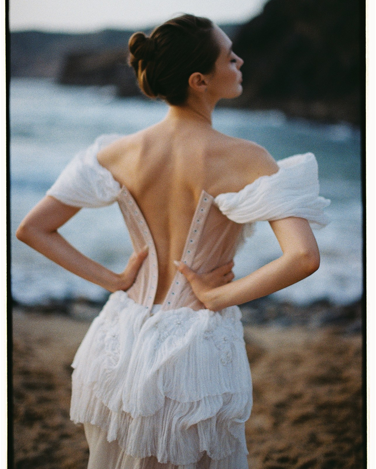 Невеста в дизайнерском свадебном платье с открытой спиной стоит у моря во время портретной фотосессии