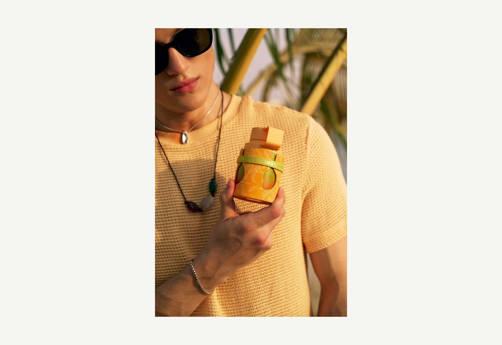 Person in sunglasses and a beige knit top holding an orange perfume bottle outdoors