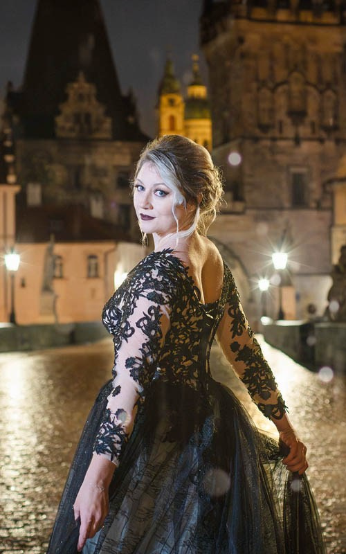 A woman wearing a black evening dress stands atop the rain-soaked Charles Bridge at night during the Gothic-inspired portrait session.