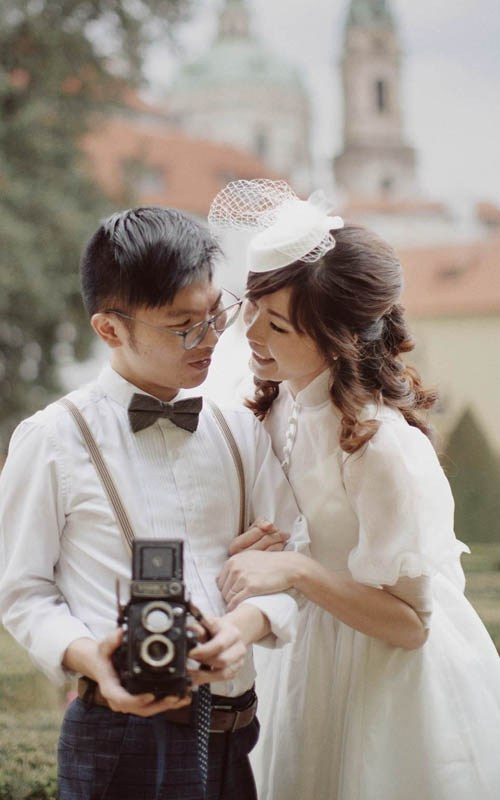 A couple dressed in vintage wedding clothes looks through the prism of a vintage camera during their Vrtba Garden pre-wedding portrait session in Prague.
