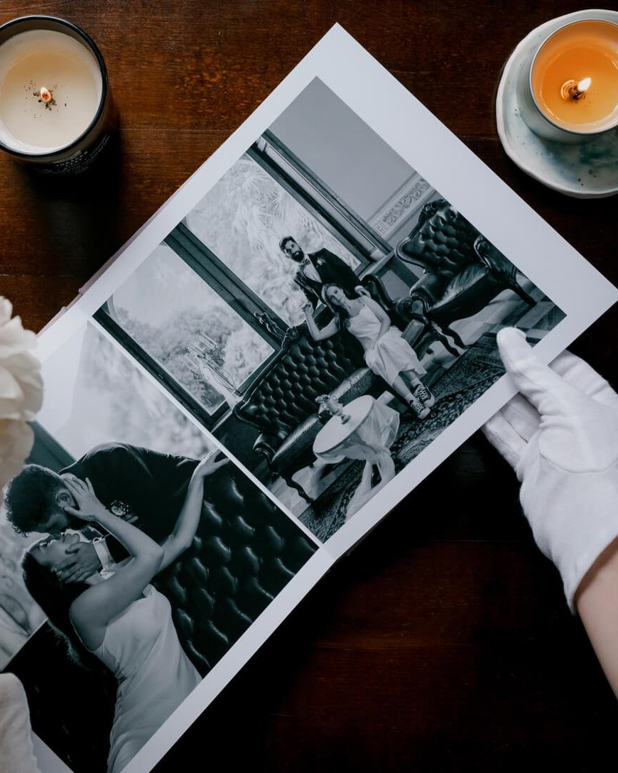 White-gloved hand turning the spread of a fine art wedding album open to intimate black-and-white portraits of a couple on a tufted leather sofa