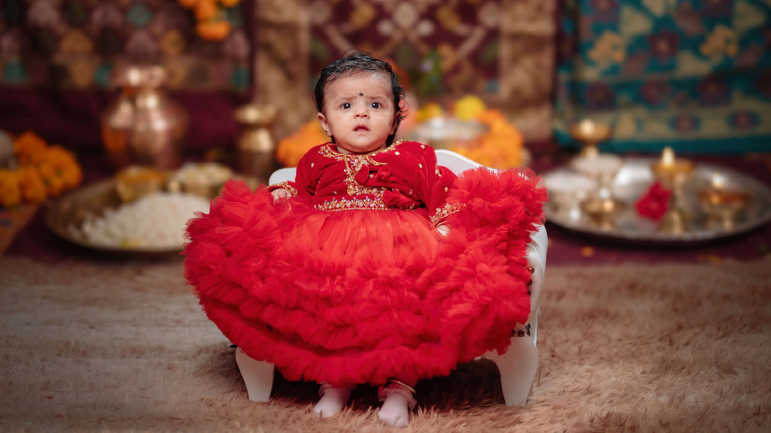 Baby in a red frilled dress sitting on a wooden floor with traditional decor in a Pasni photoshoot
