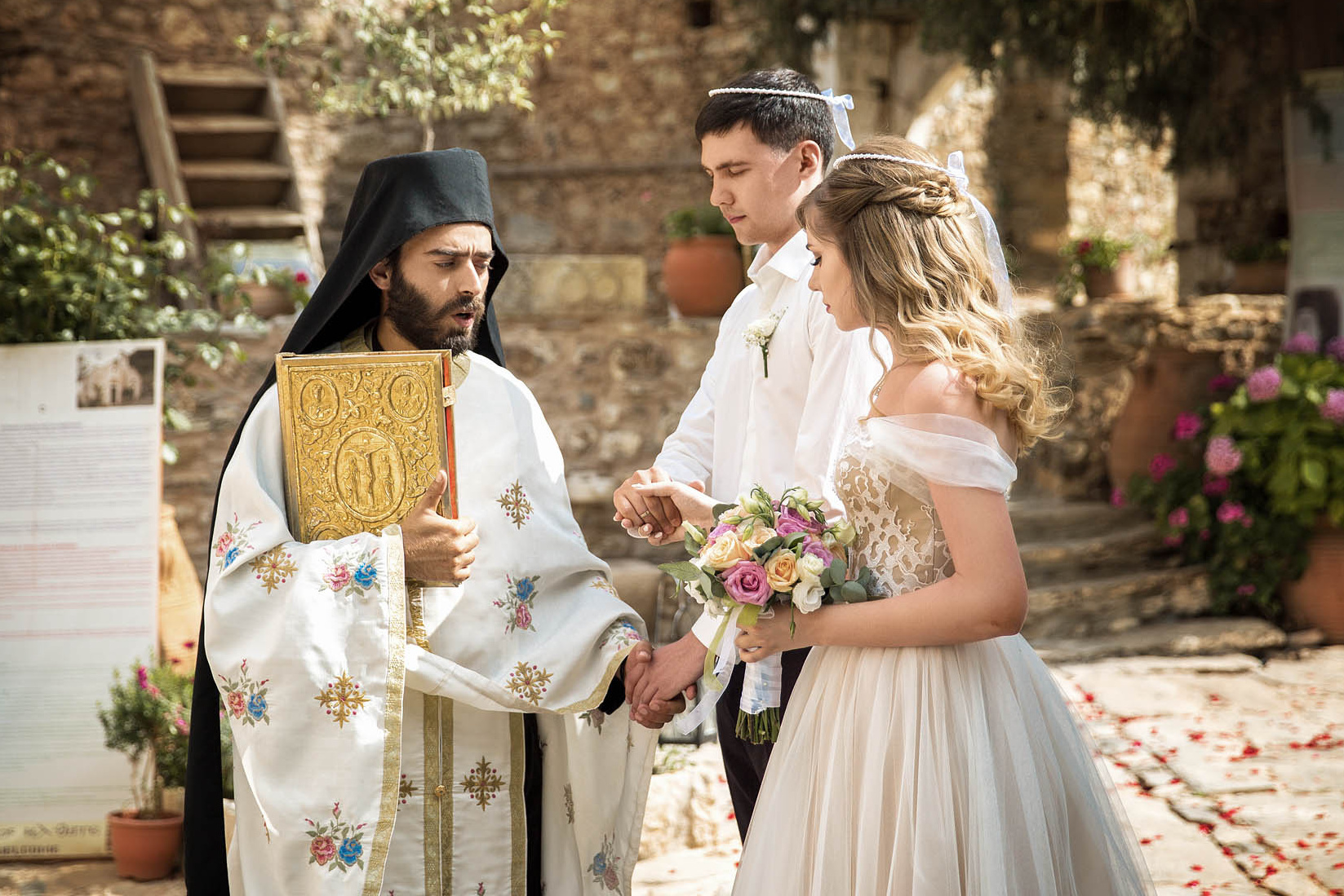 Святой отец держит за руки венчающуюся пару во время таинства на Крите. Фотограф, свадьба в Греции.