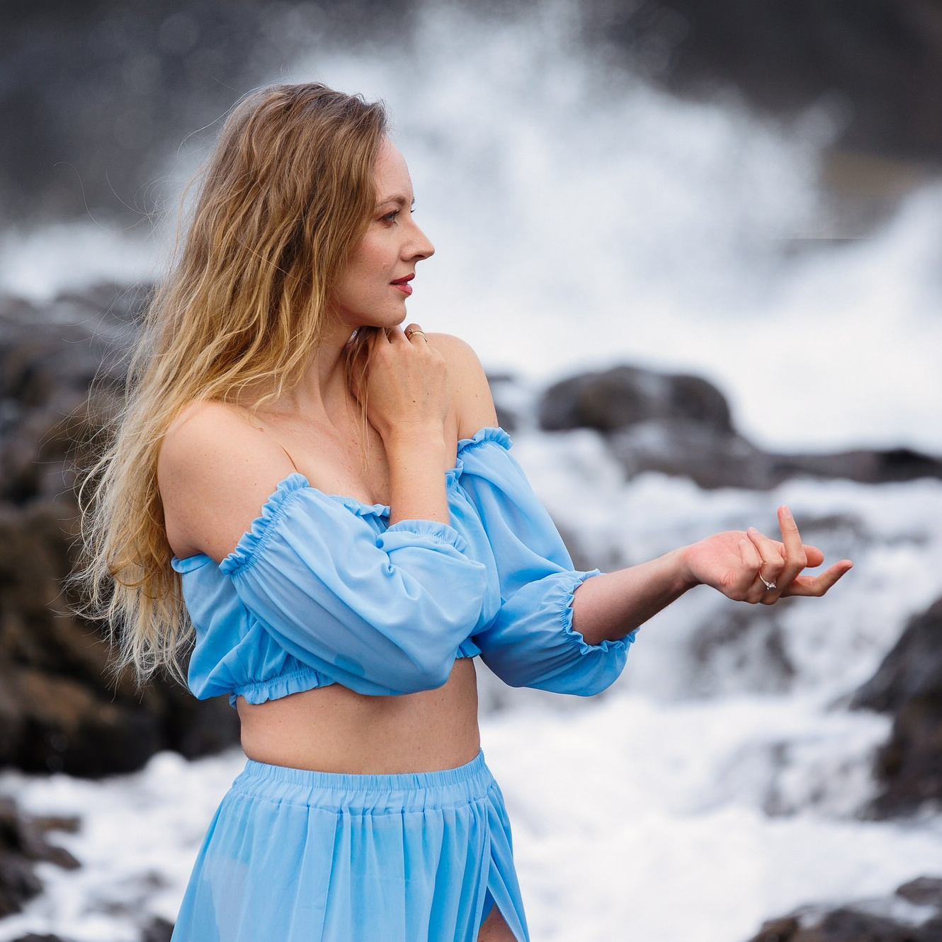 Blonde woman in blue off-shoulder top in profile, ocean waves crashing behind, Gran Canaria coast