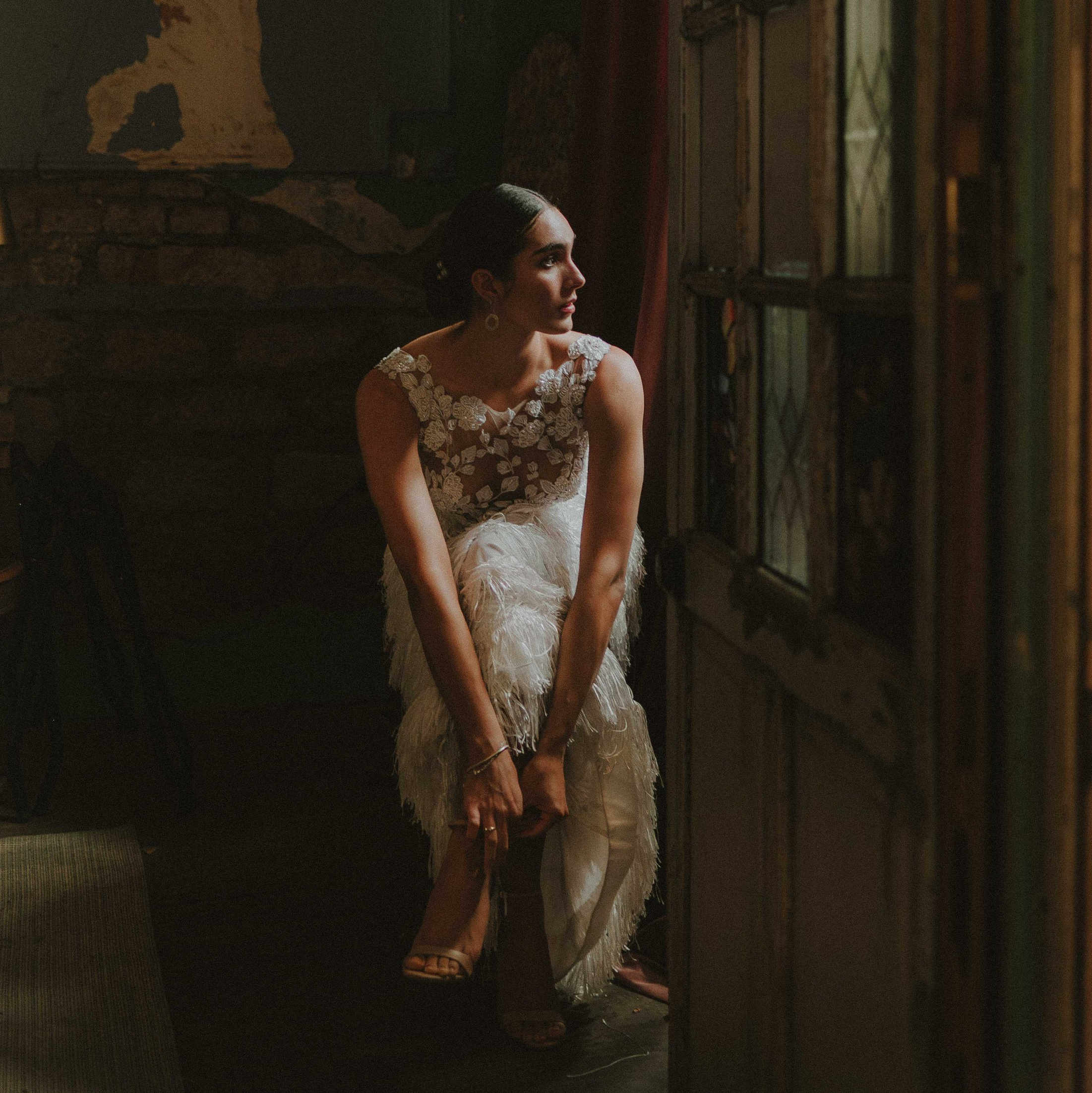 A candid bridal preparation scene showing a bride in an elegant sleeveless lace and feather wedding gown, seated near a rustic wooden and glass door. Soft natural light streams through the vintage window, illuminating her profile as she leans forward to adjust the strap of her nude high-heeled shoes. The textured walls, peeling paint, and exposed stone create a romantic, old-world atmosphere, perfect for intimate wedding photography in a historic venue
