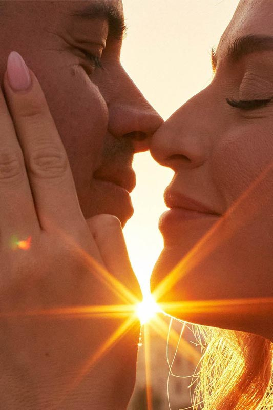 A gorgeous couple lovingly embraces as the sun flares vibrantly between their faces.