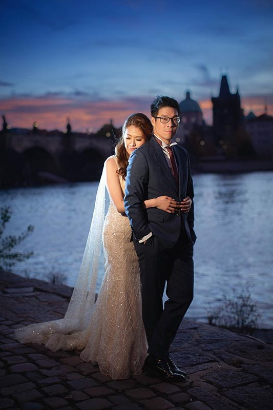 A stylish American couple dressed in evening attire embrace near the bank of the Vltava River as the sky flares above the Charles Bridge in Prague.