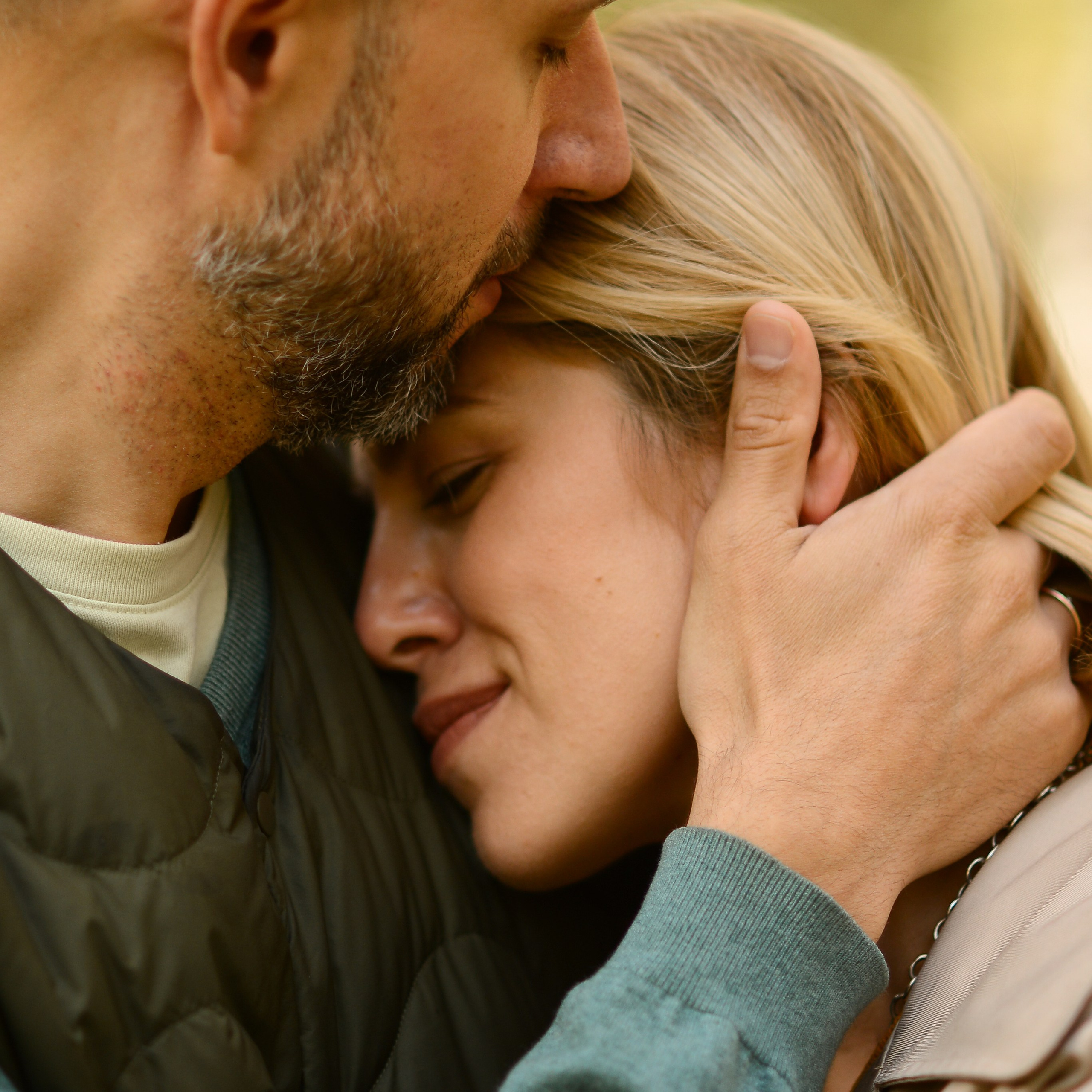 Man gently embracing a woman, resting his forehead on her head — a quiet, intimate moment of love captured in soft autumn light.