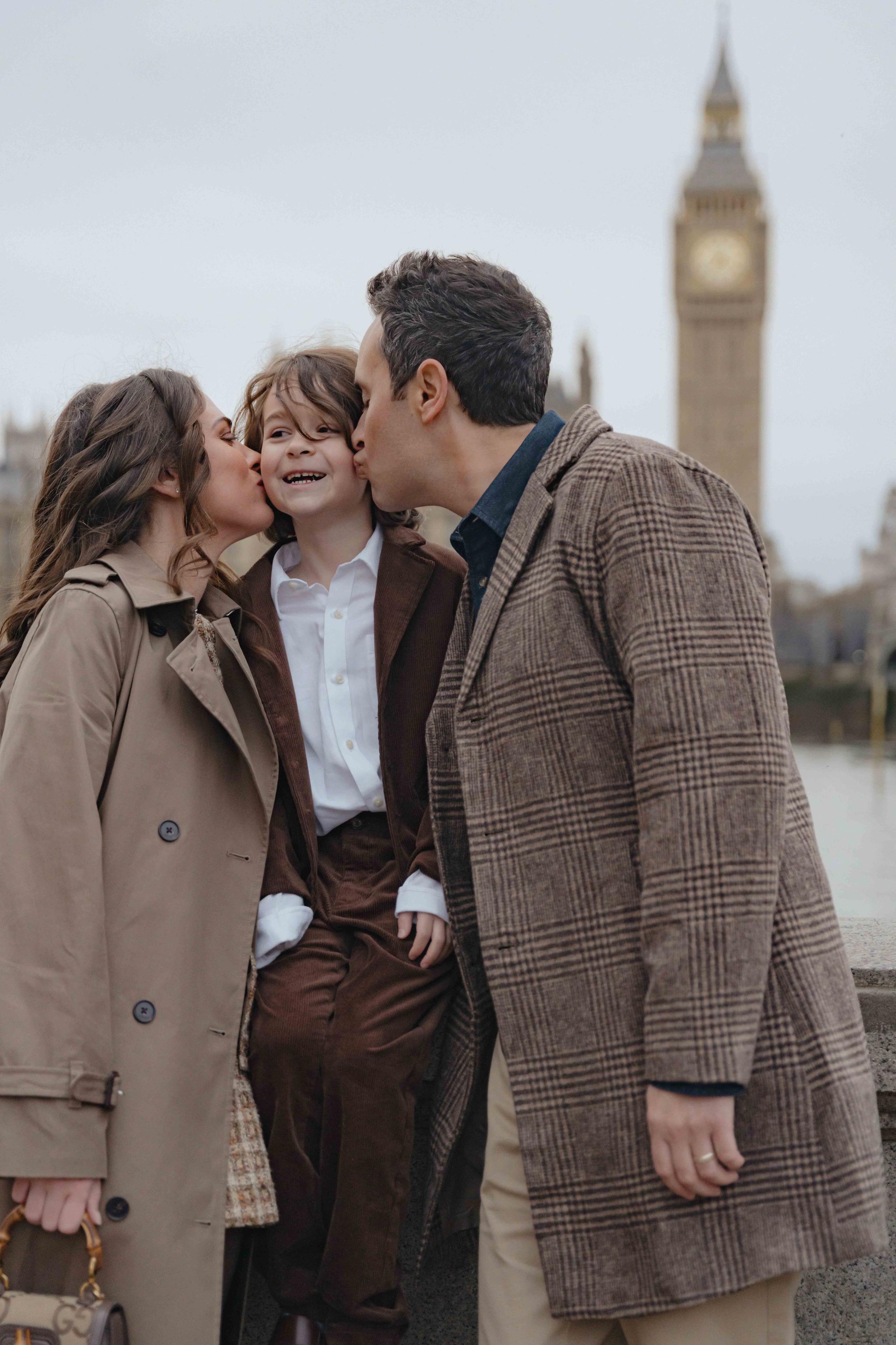 Parents kissing child during London family photoshoot at Westminster Bridge with Big Ben view.