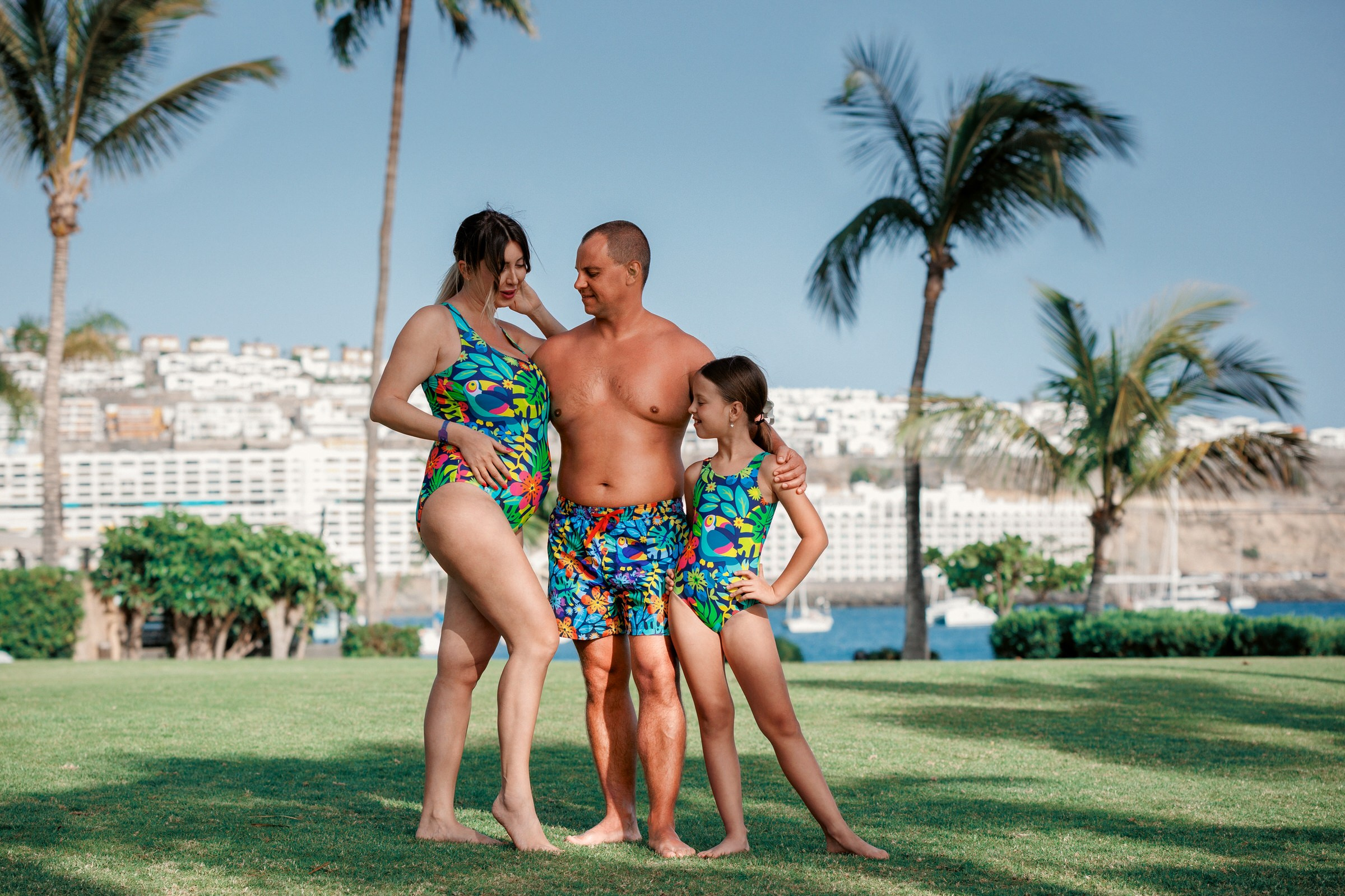 Anfi beach Gran Canaria A family of three wearing matching tropical swimsuits stands together on grass with palm trees and buildings in the background.