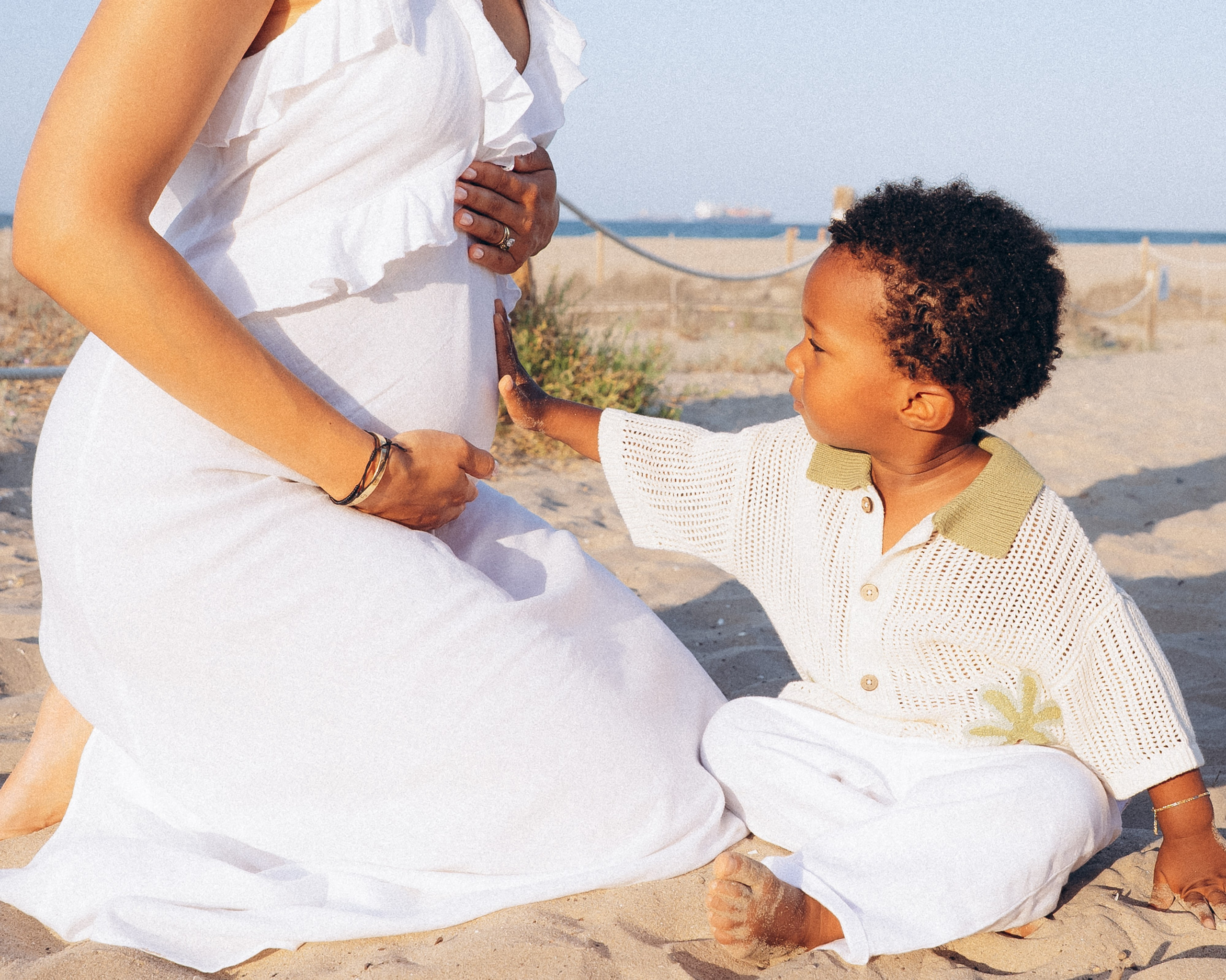 Maternity photoshoot in Valencia, Spain, capturing a tender moment as a young child gently touches his mother’s pregnant belly on a sandy beach, photographed in soft natural light, highlighting family connection and pregnancy emotions by the Mediterranean Sea.