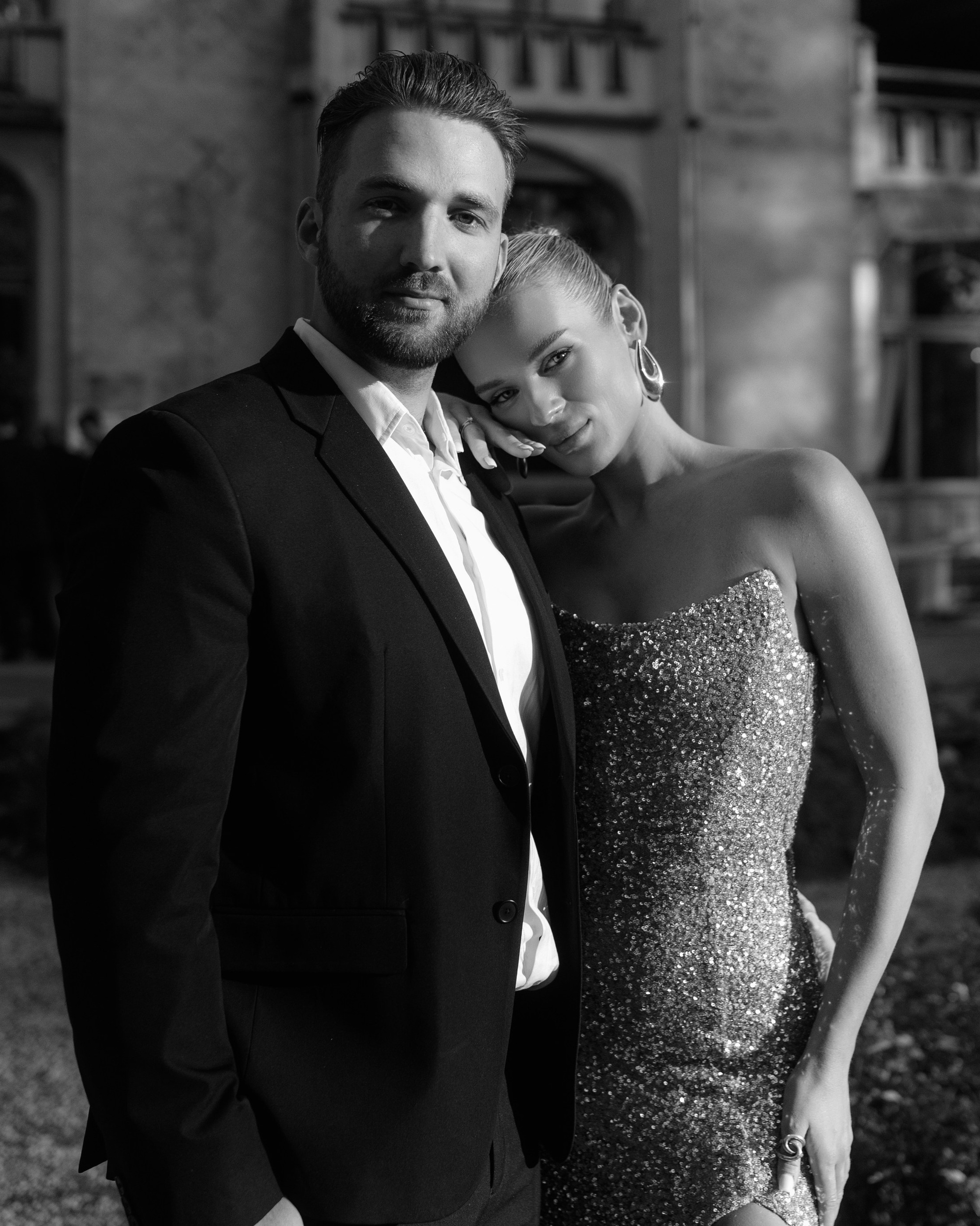 Black and white photo of a couple on the beach. A man in a light shirt gently hugs a woman in a long slit dress, they look calm and happy against the background of the sea and the horizon