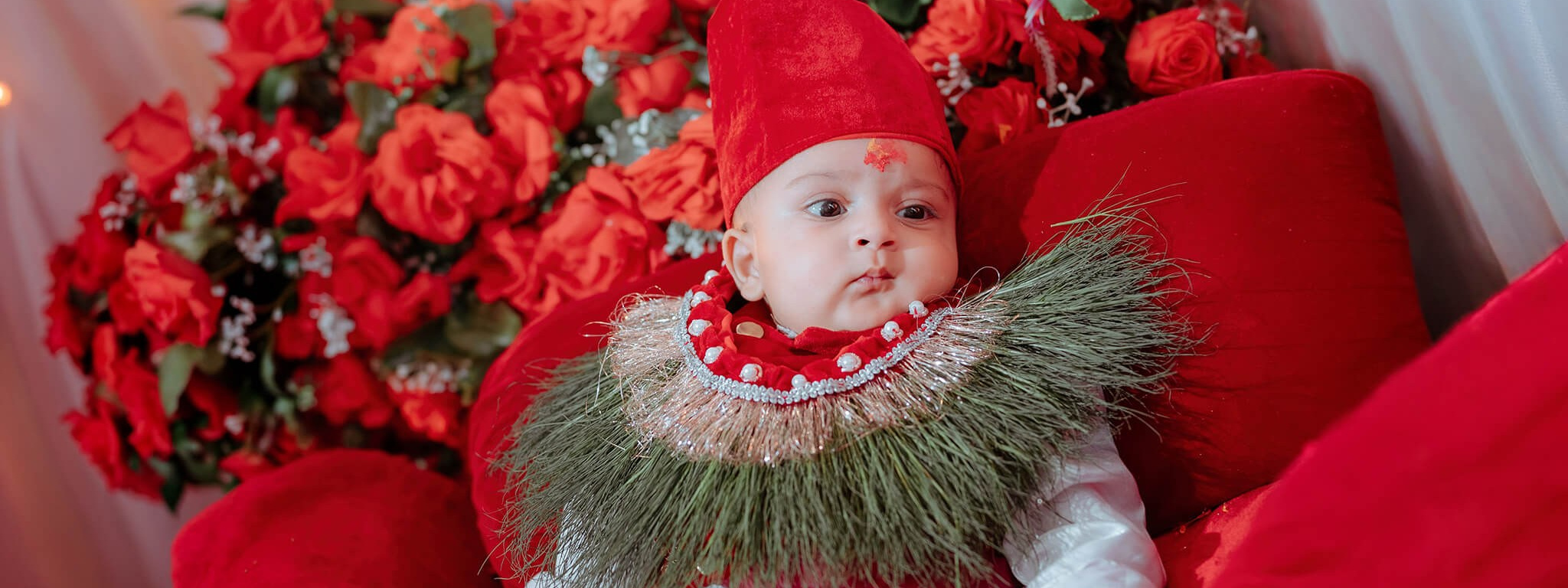Baby sitting among red flowers and cushions in a Pasni photoshoot setup