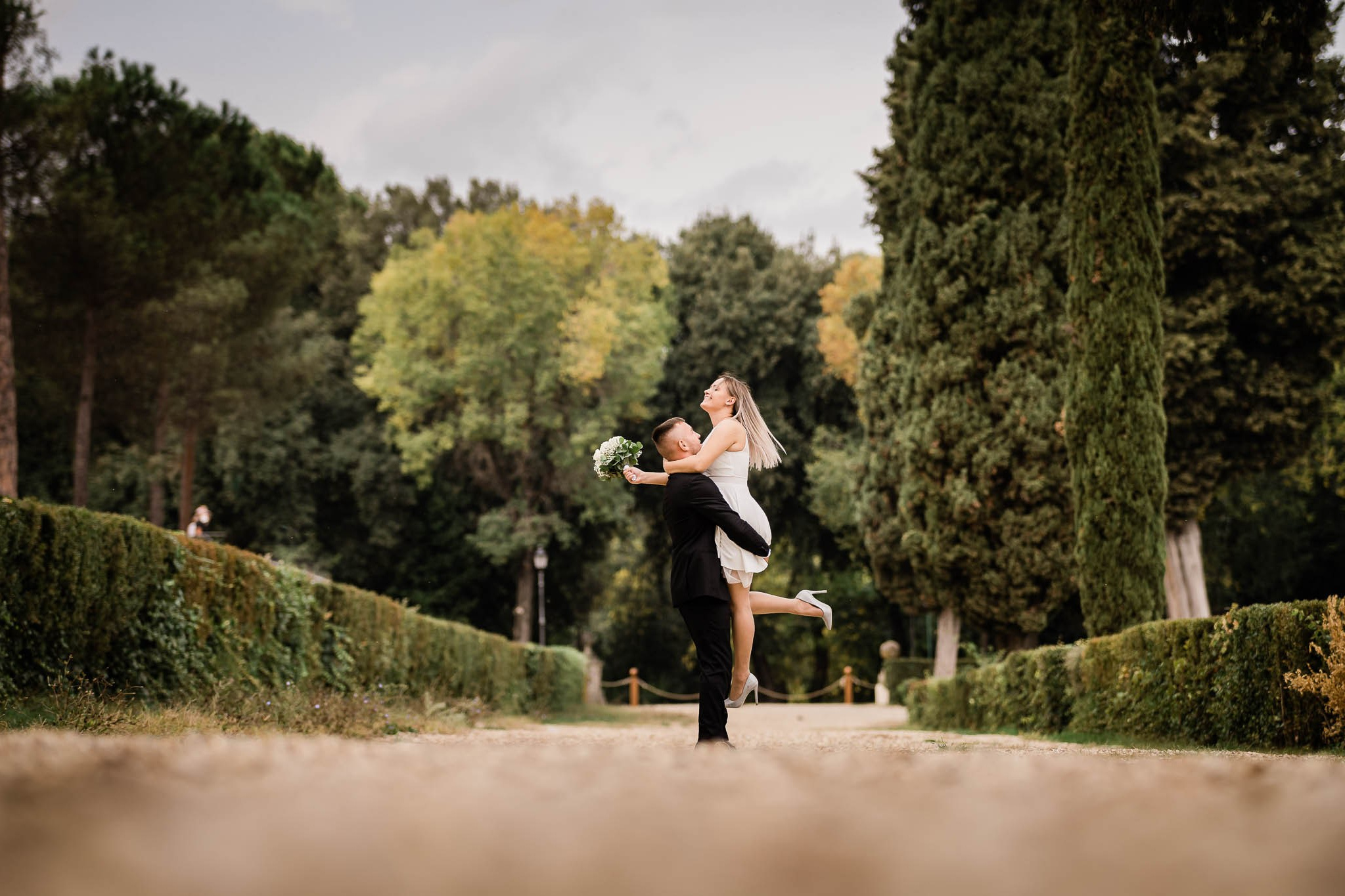 Gioioso sposo solleva la sua sposa in un abbraccio mentre danzano su un viale alberato, circondati dalla bellezza naturale di un parco, catturando un momento di pura felicità e celebrazione durante un servizio fotografico prematrimoniale.