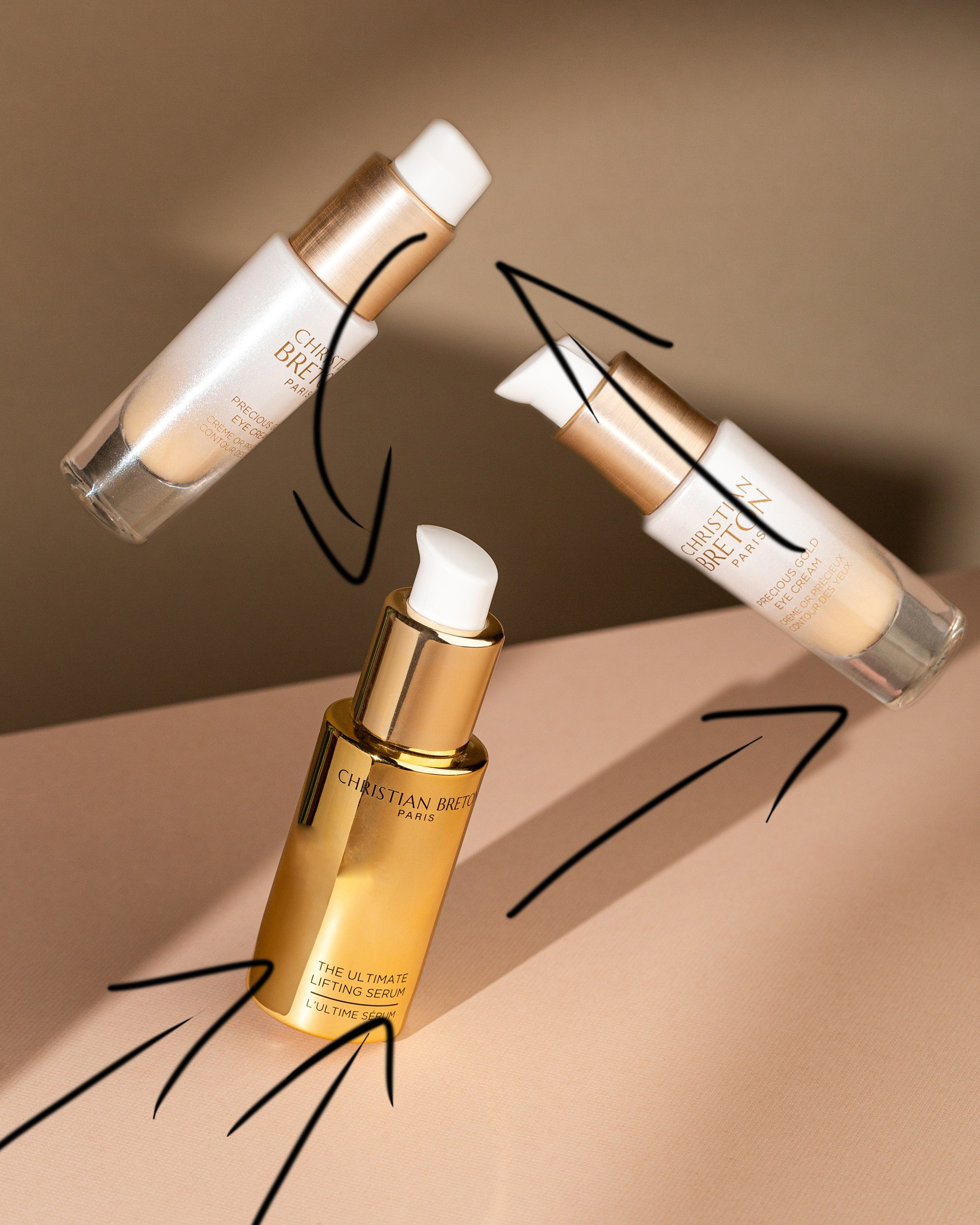 Christian Breton Paris kosmetik məhsulları – dinamik kompozisiyanın hərəkət istiqamətlərini göstərən qara xətlərlə işarələnmiş foto; mərkəzdə qızılı The Ultimate Lifting Serum, ətrafda isə iki Precious Gold Eye Cream şüşəsi.