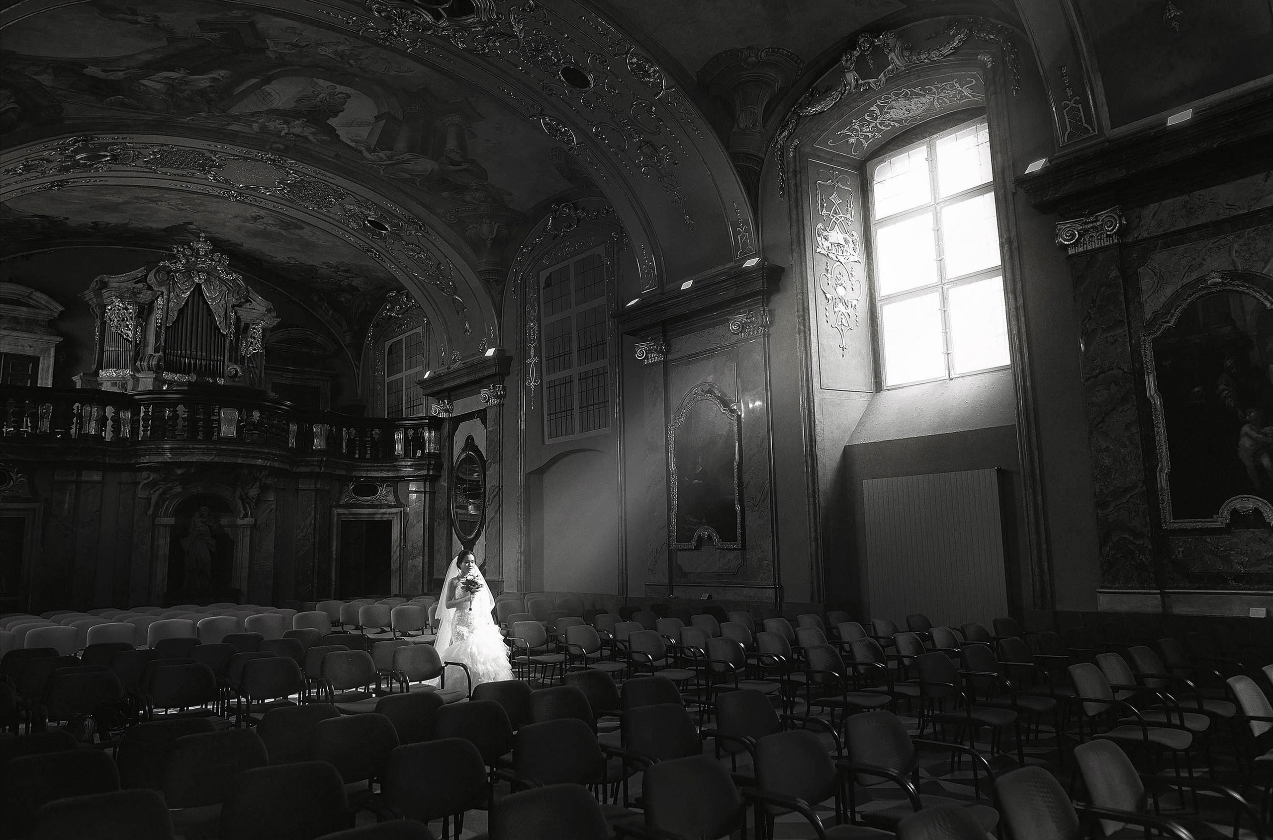 An asian bride walks into a shaft of sunlight that is filtering through a window in the Mirror Chapel located in the Klementinum in Prague.