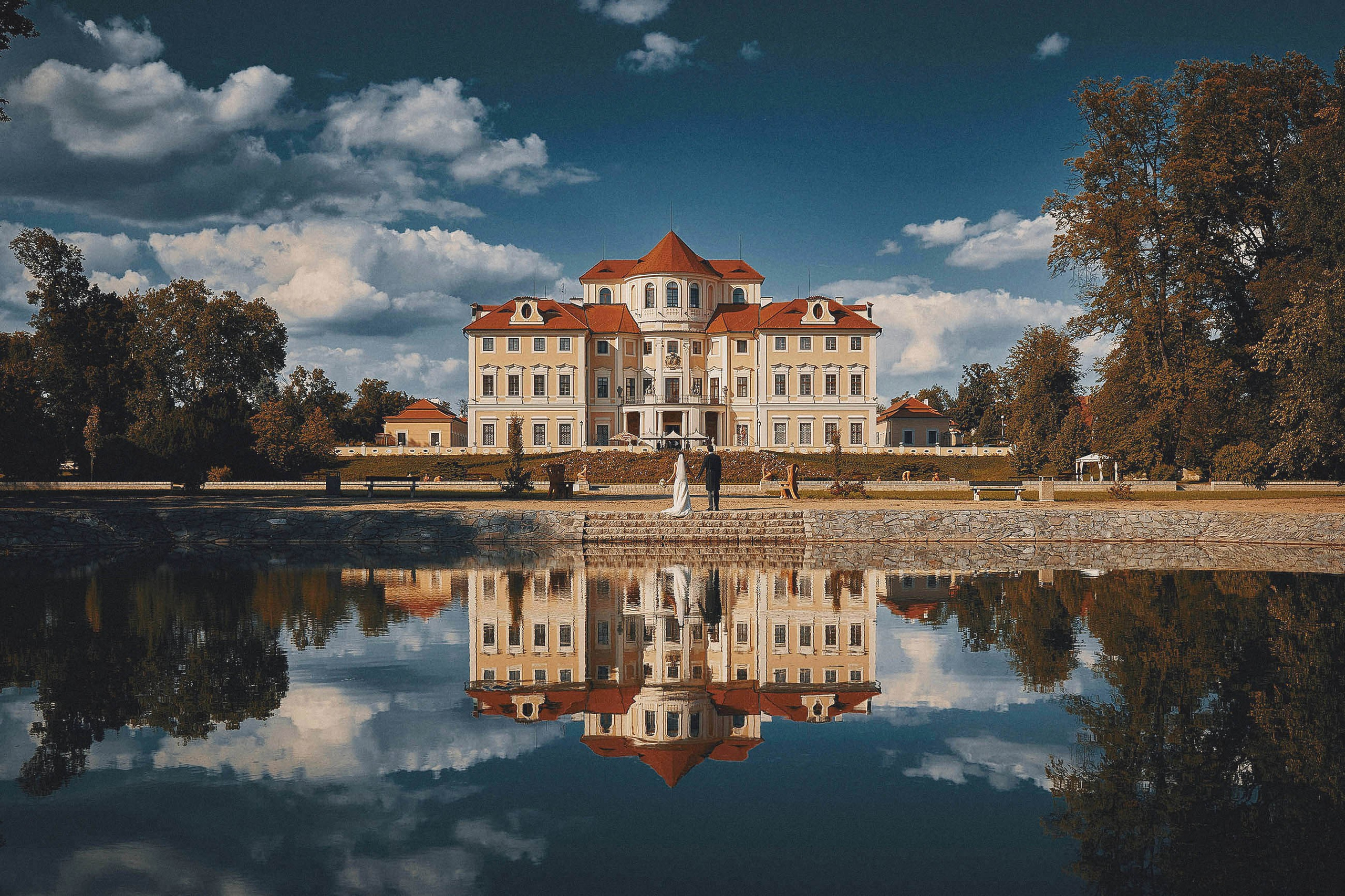 Newlyweds facing stately chateau with vibrant blue sky reflected in waters, Bohemia wedding portrait