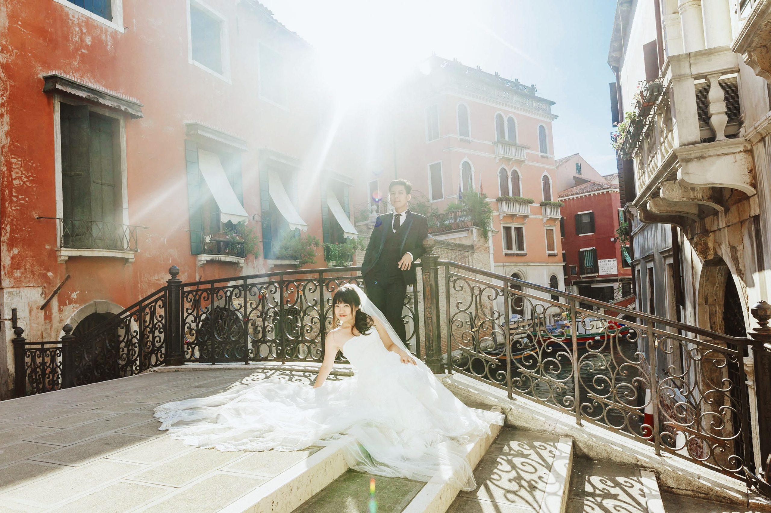 Stylish Asian newlyweds pose for a portrait atop a small bridge in Venice, Italy, as the early morning sunlight bathes them in light - Kurt Vinion Photography