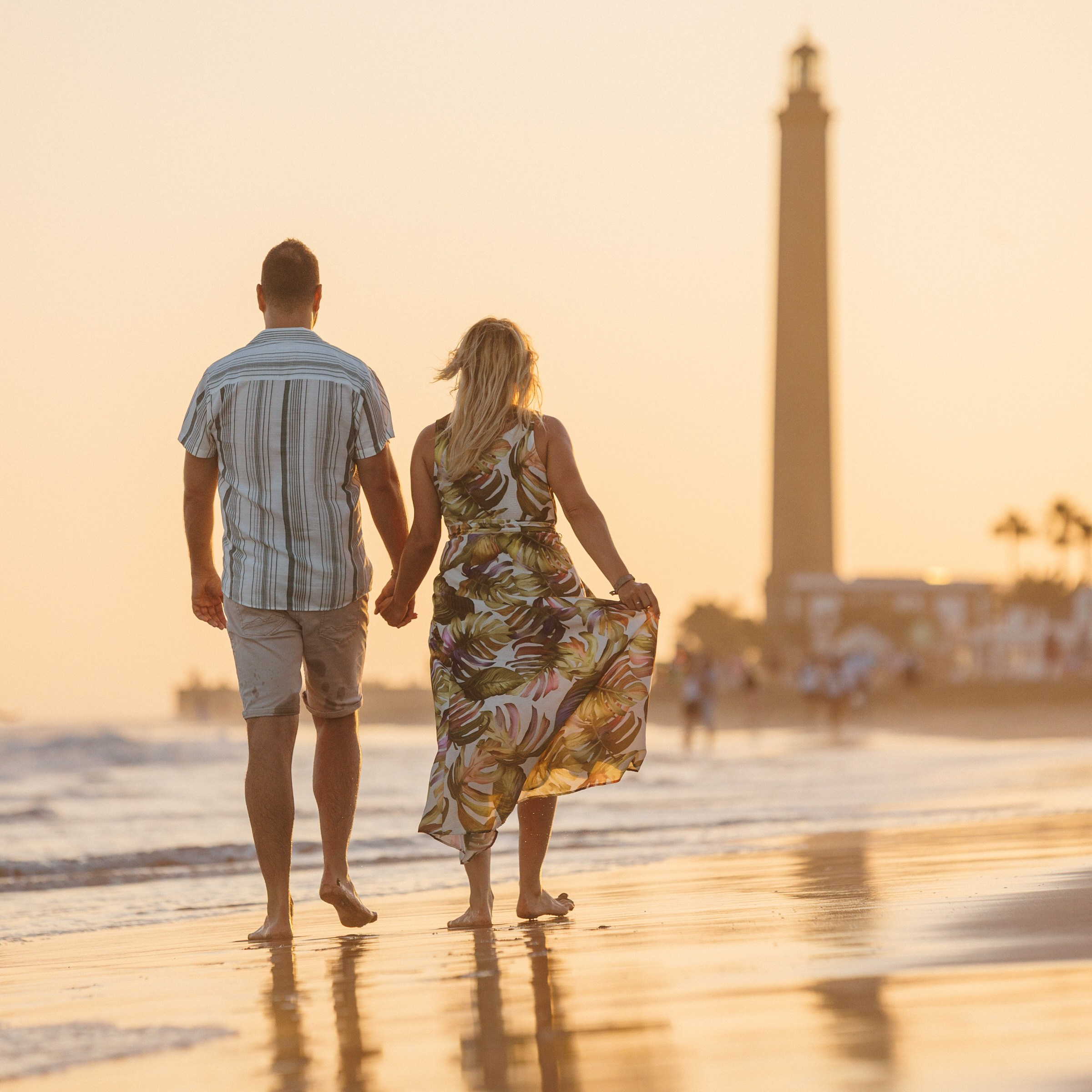 A man and woman walk hand in hand along a beach at sunset with a tall lighthouse in the background Faro Maspalomas Photographer