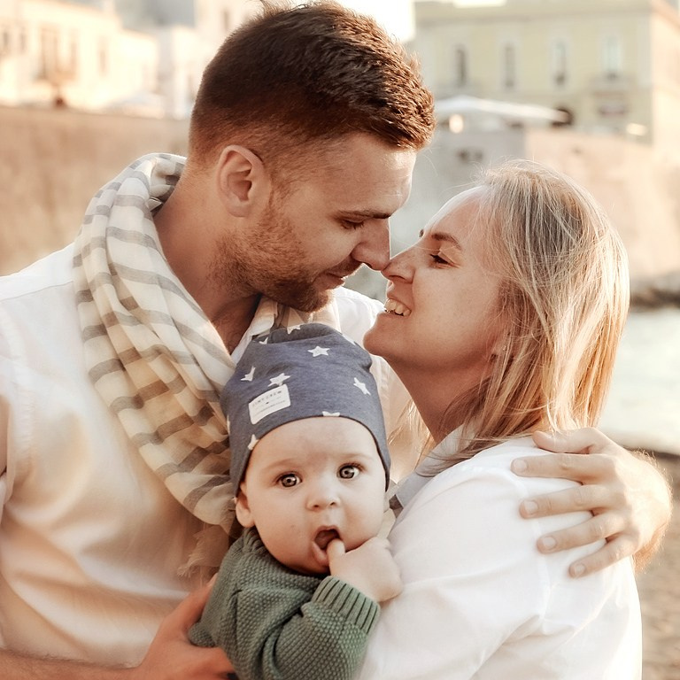Young parents smiling as they hold their baby close — warm family moment during a sunset stroll in Polignano a Mare