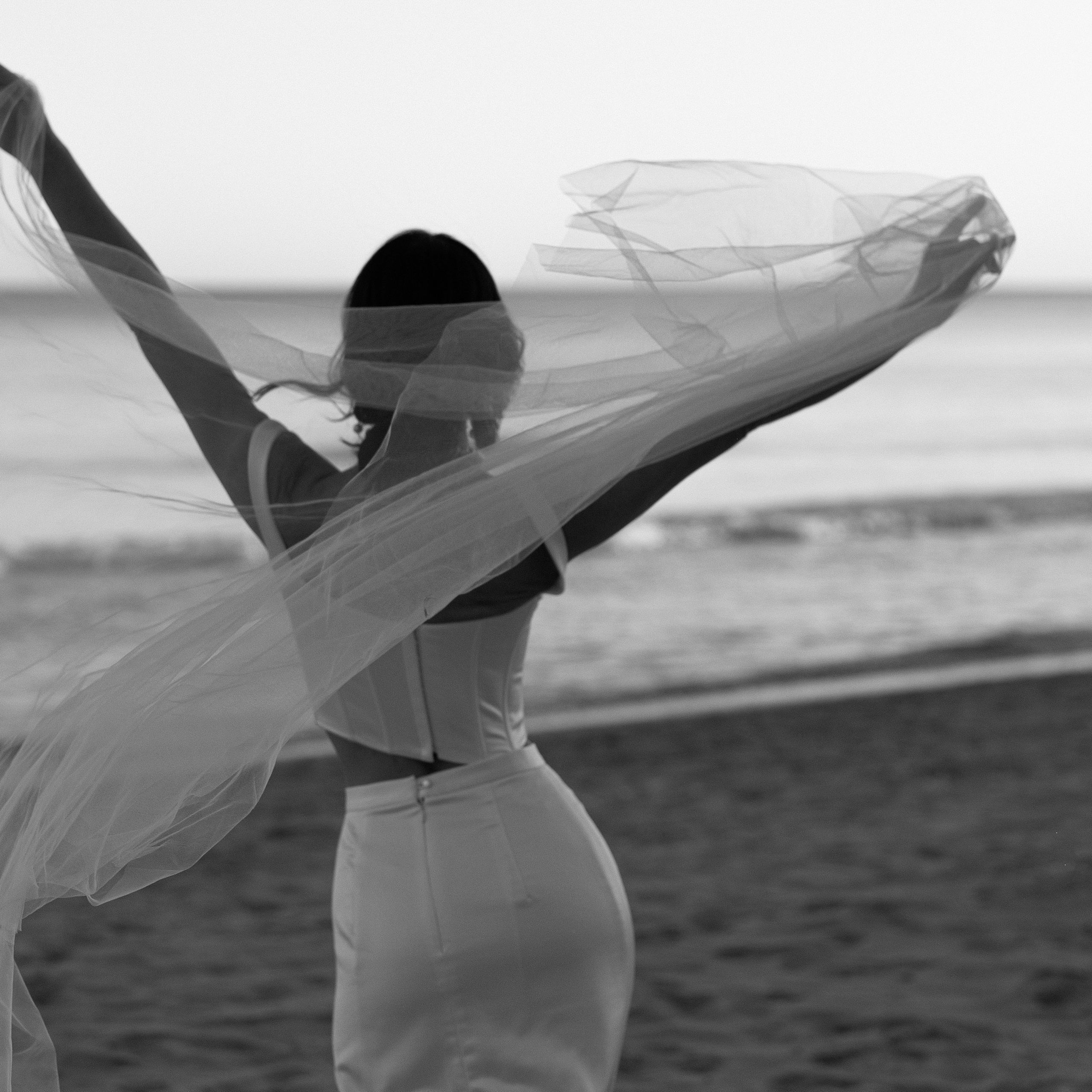 A black-and-white photo of a bride standing on the beach with her back turned, holding her veil in the air as it flows in the wind, with the ocean in the background.