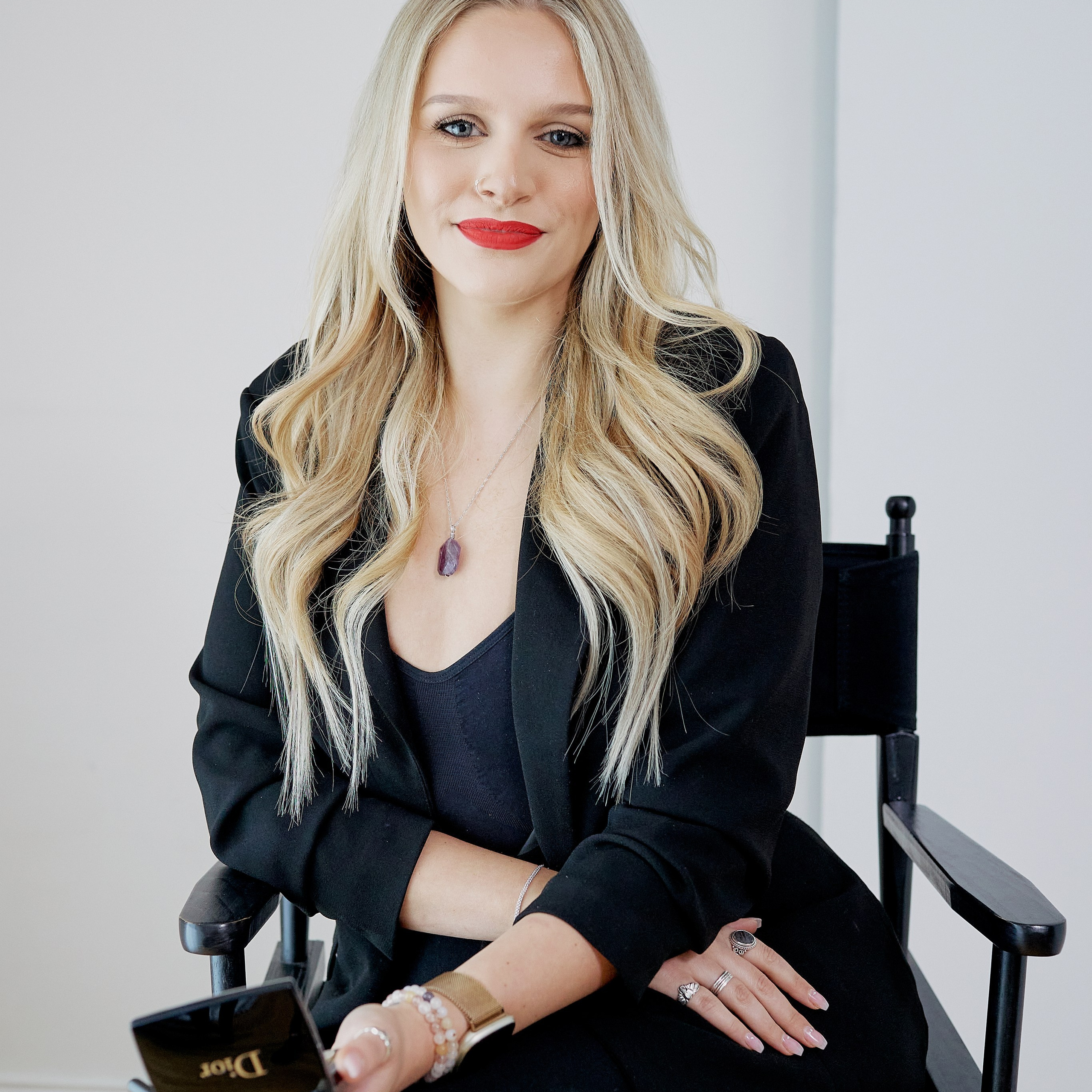 HEADSHOT OF A WOMAN IN BLACK SITTING ON A CHAIR HOLDING MAKEUP IN HER HAND