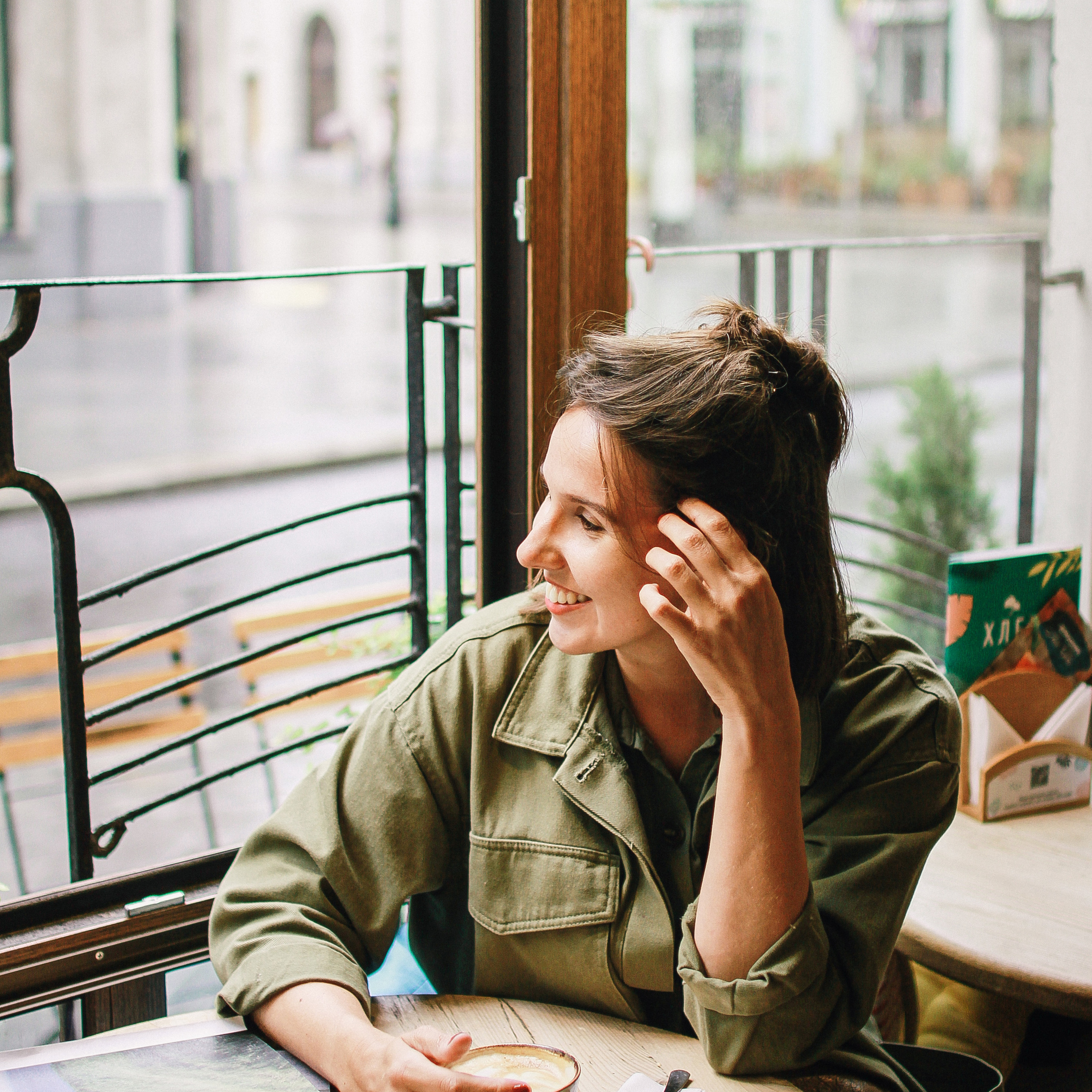 Девушки в куртке цвета хаки, сидит у окна в кафе, улыбается и держит кружку с кофе одной рукой