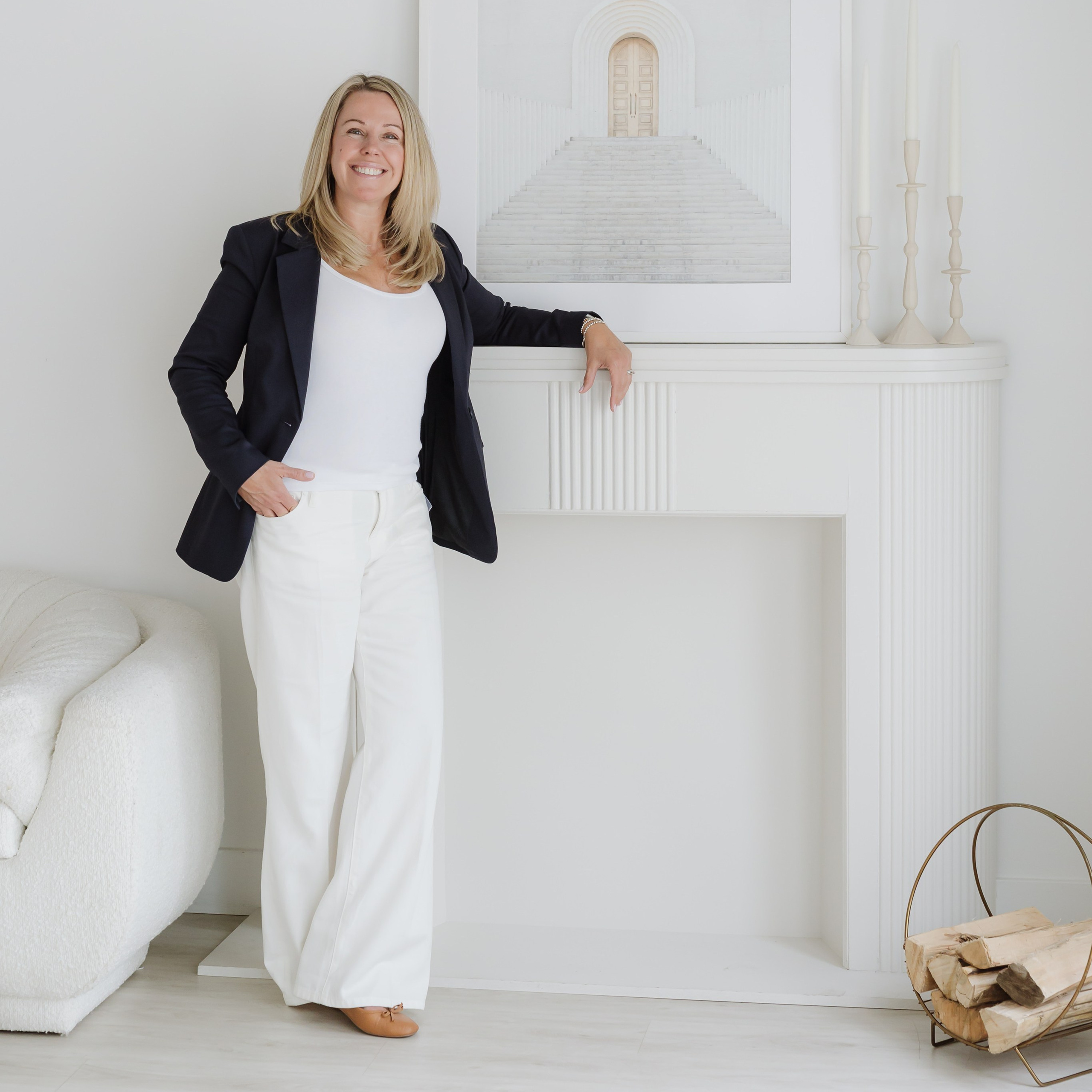 PERSONAL BRANDING PHOTO OF WOMAN DRESSED IN WHITE WITH BLACK BLAZER, LEANING ON A WHITE FIREPLACE