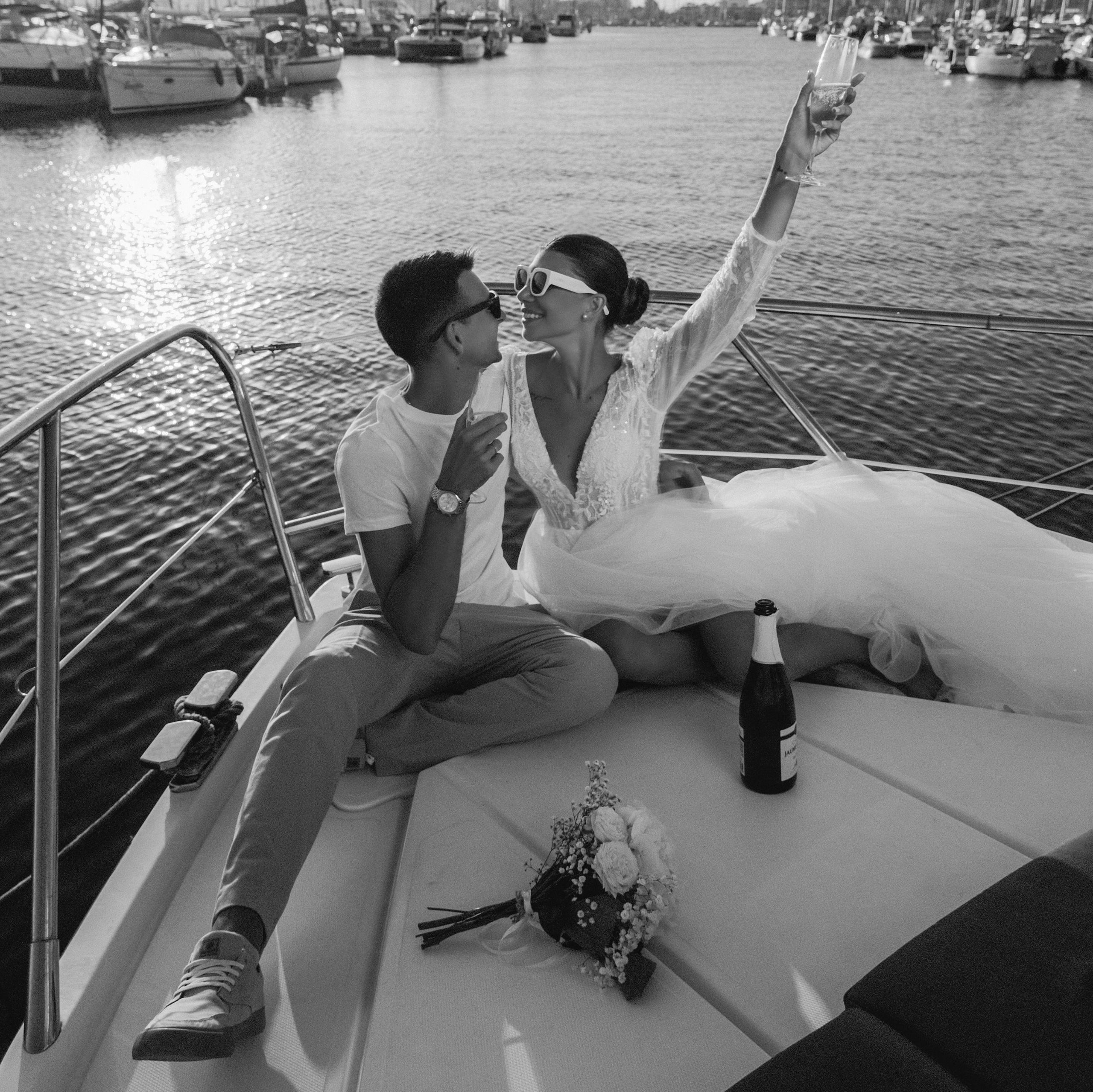 A bride and groom sitting on a boat, celebrating with champagne as the bride raises her glass. The marina with boats and reflections on the water creates a romantic backdrop.