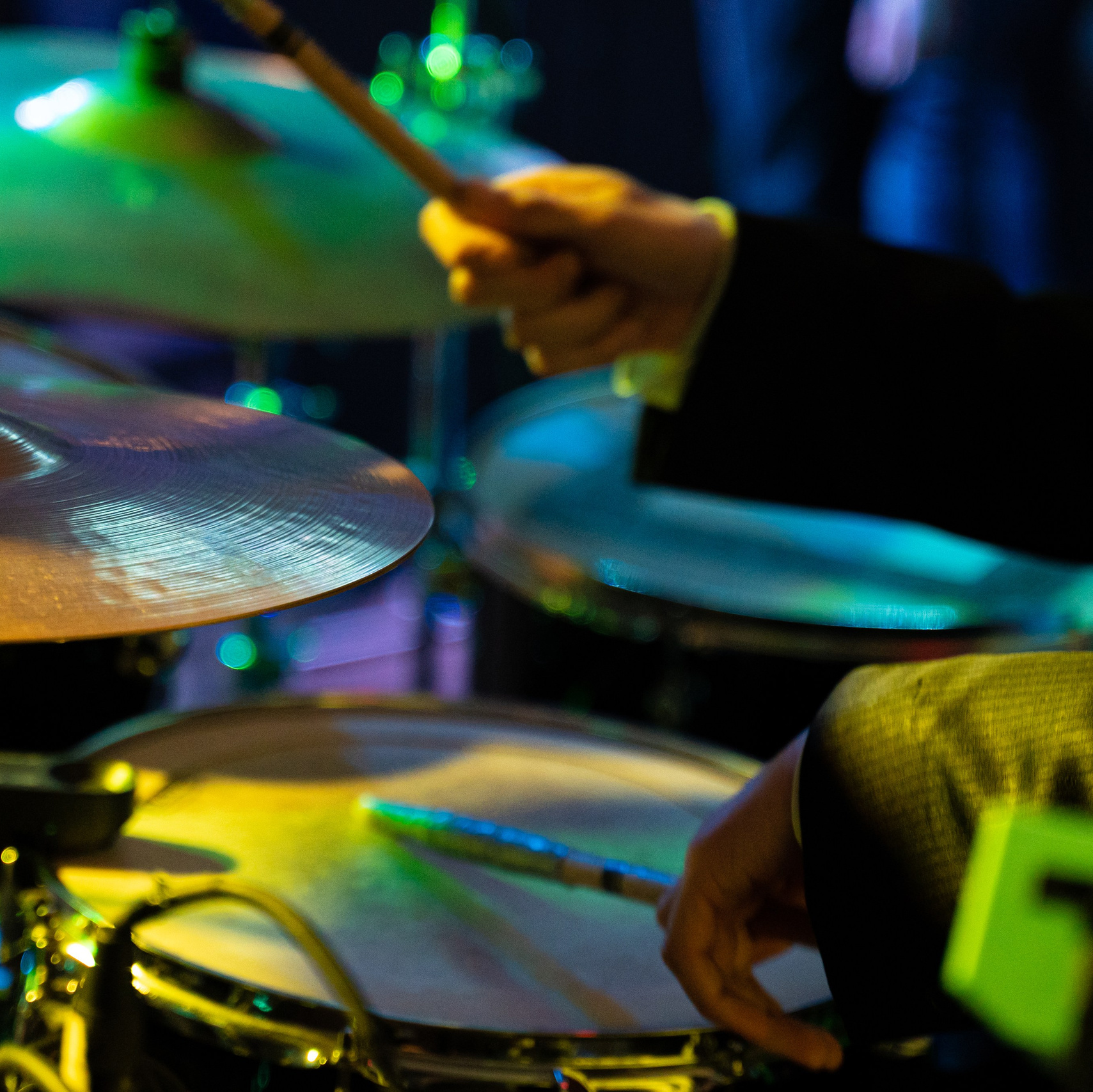 Detaliu dinamic cu mâinile unui toboșar în timpul unui concert live, lumini de scenă colorate, fotografie de eveniment realizată de incadru.ro.